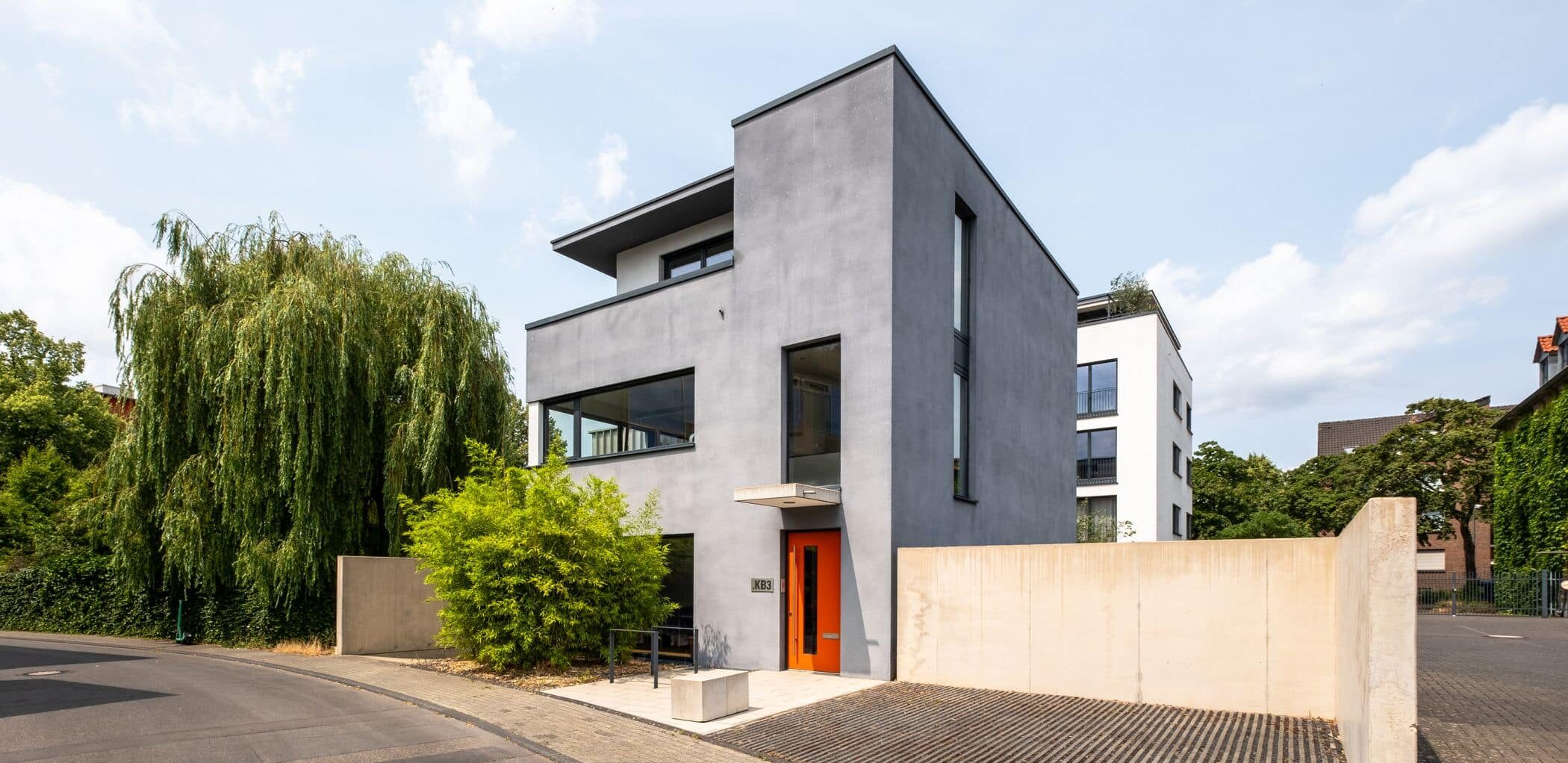 Ein modernes, dreistöckiges Haus mit glatter grauer Fassade und großen Fenstern steht auf einem Eckgrundstück neben einer gepflasterten Straße. Perfekt für alle, die immobilien köln suchen. Es hat eine orangefarbene Eingangstür, grüne Bäume, Sträucher und eine minimalistische Einfahrtsmauer.