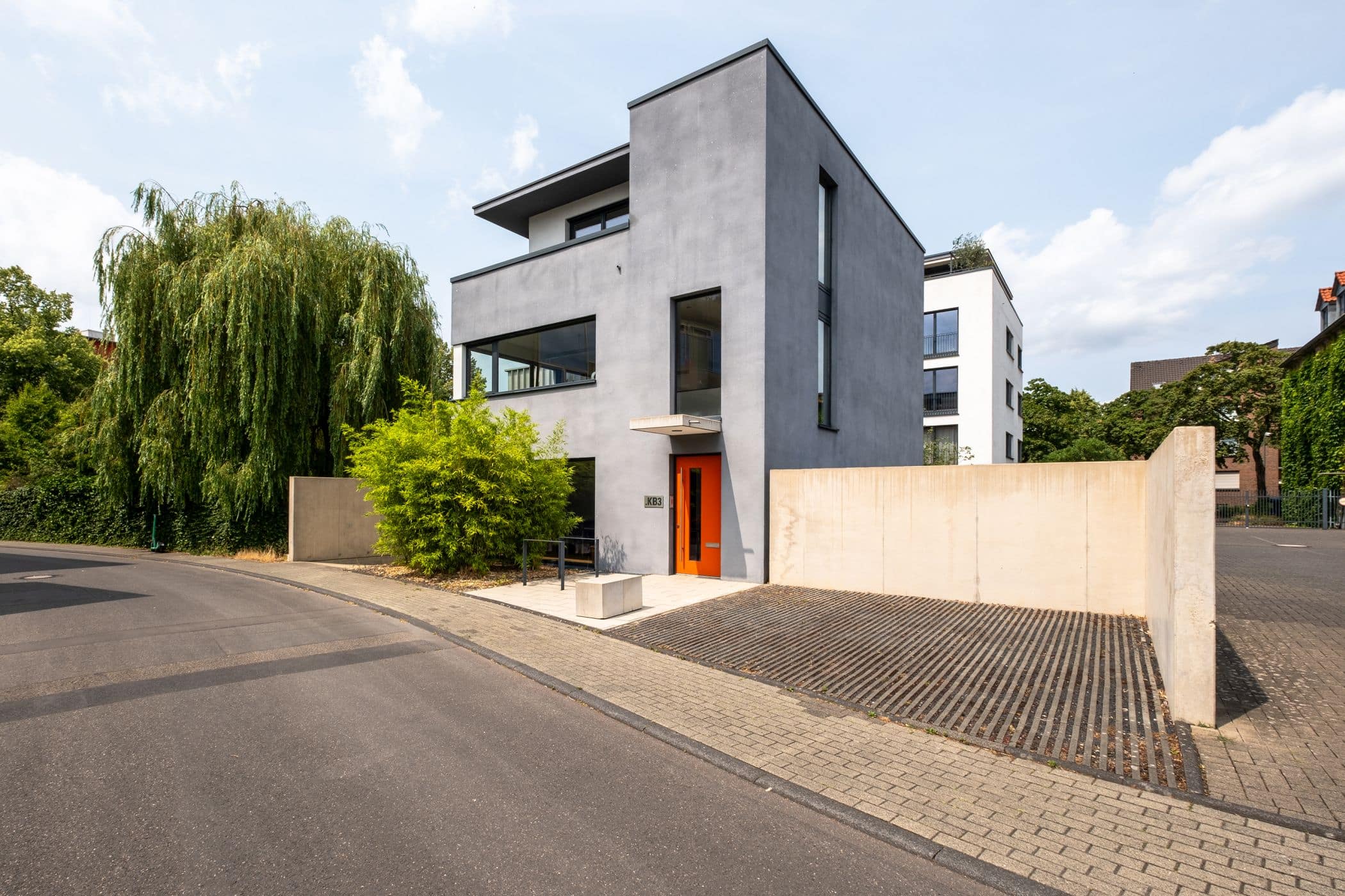 Ein modernes, dreistöckiges Haus mit glatter grauer Fassade und großen Fenstern steht auf einem Eckgrundstück neben einer gepflasterten Straße. Perfekt für alle, die immobilien köln suchen. Es hat eine orangefarbene Eingangstür, grüne Bäume, Sträucher und eine minimalistische Einfahrtsmauer.