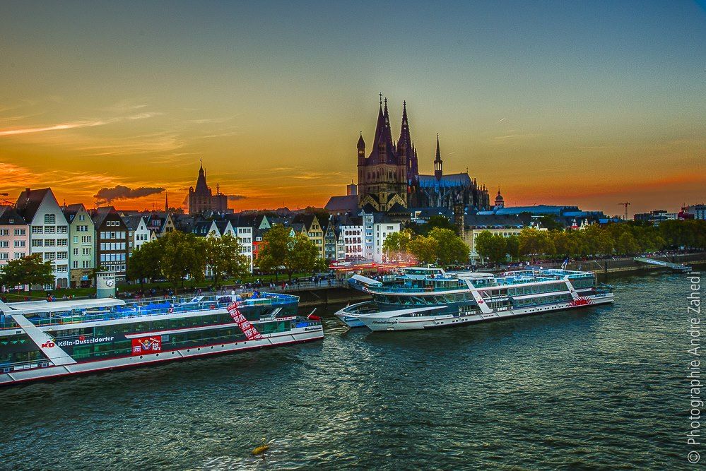 Farbenfroher Sonnenuntergang über dem Rhein in Köln, Deutschland, mit großen Booten, die am Ufer angedockt sind, und dem Kölner Dom prominent im Hintergrund.