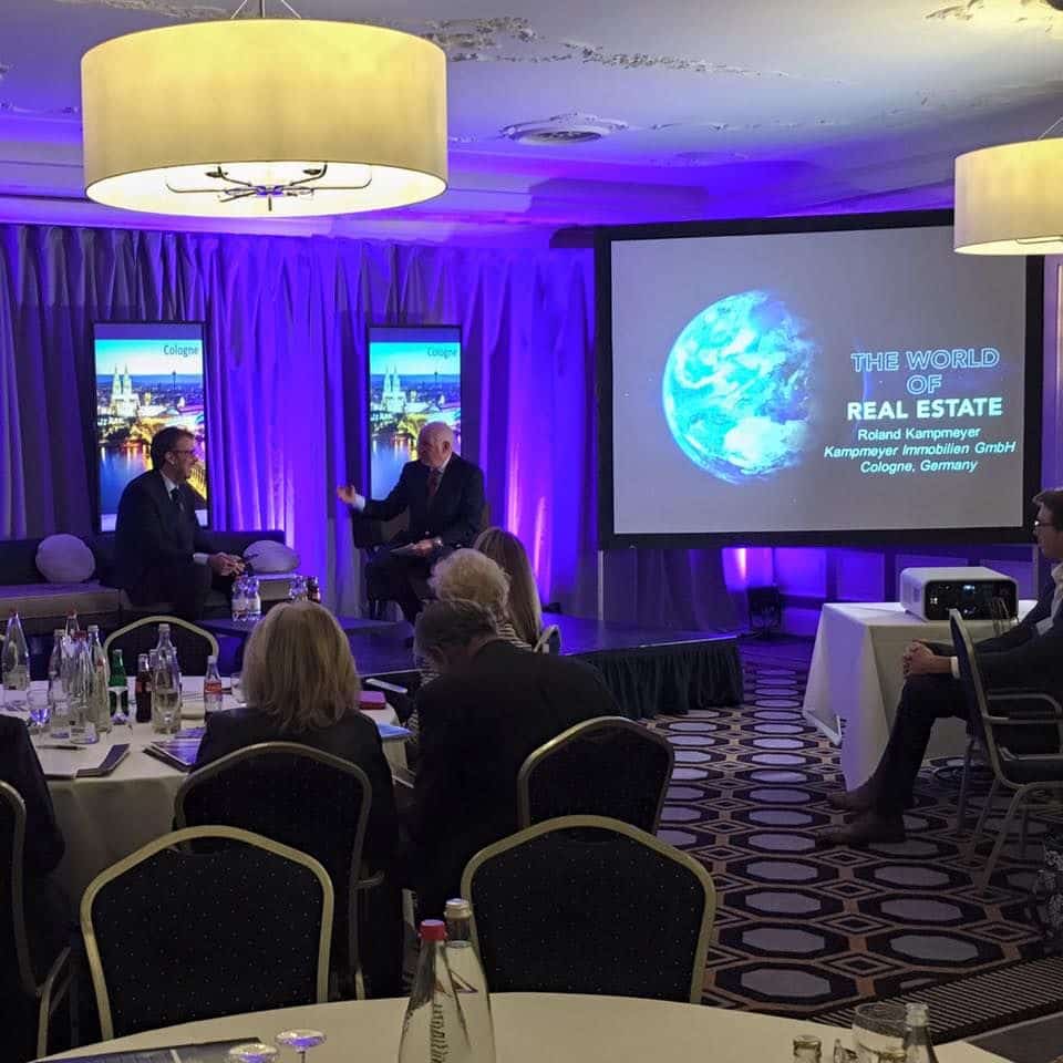 Zwei Männer diskutieren auf der Bühne einer Konferenz in einem Bankettsaal eines Hotels. Auf einem Bildschirm hinter ihnen wird "The World of Real Estate" mit einem Bild der Erde angezeigt. Die Anwesenden sitzen an runden Tischen.