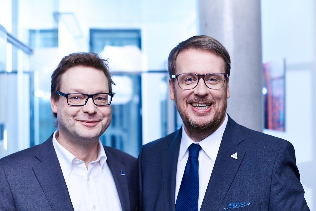 Zwei Männer in Geschäftsanzügen und mit Brillen stehen nebeneinander und lächeln in die Kamera in einer modernen, hellen Büroumgebung mit einem blauen Farbton.