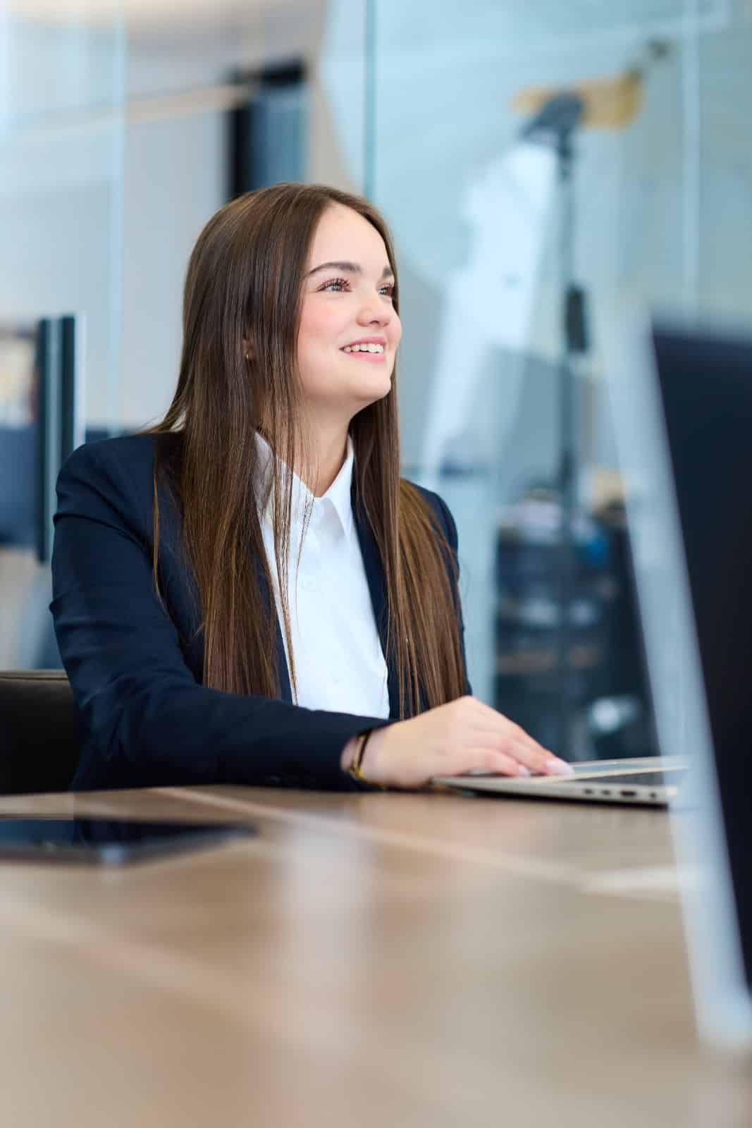 Eine junge Frau in Business-Kleidung, möglicherweise eine Immobilienmaklerin aus Köln-Porz, sitzt an einem Schreibtisch in einem modernen Büro, lächelt und arbeitet an einem Laptop; der Hintergrund ist unscharf, und auf dem Tisch ist ein weiteres Gerät zu sehen.