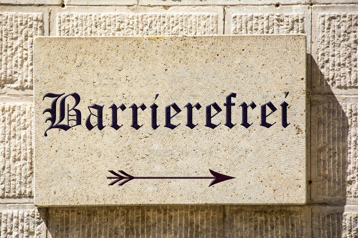 Ein steinernes Schild an einer strukturierten Wand trägt die Aufschrift Barrierefrei in schwarzer Schrift und weist mit einem Pfeil nach rechts auf einen zugänglichen oder barrierefreien Weg hin.