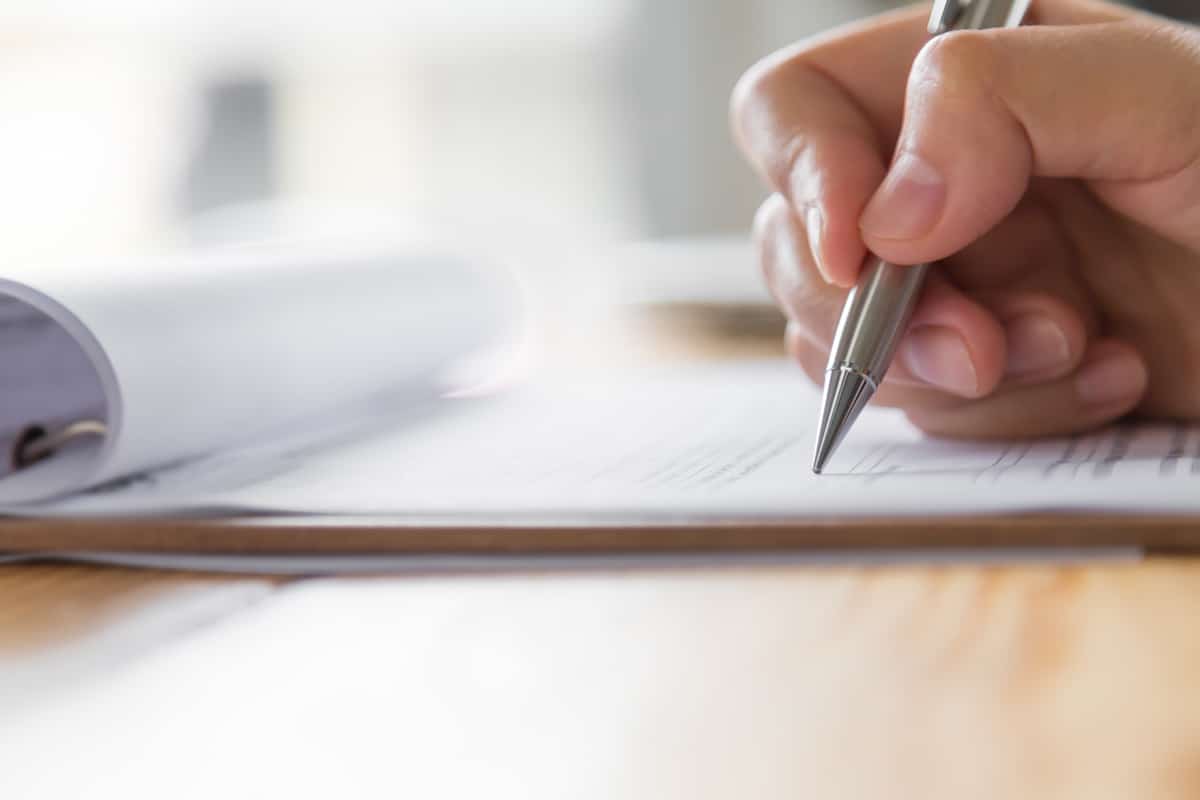 Nahaufnahme einer Hand, die einen Stift hält und auf ein Stück Papier schreibt, mit einem aufgerollten Dokument im Hintergrund auf einer Holzfläche.