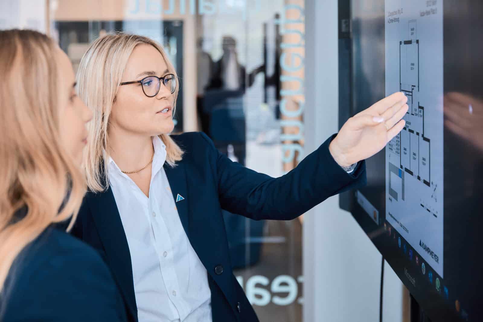 Zwei Frauen in Business-Kleidung stehen vor einem Touchscreen; eine von ihnen – eine erfahrene Immobilienmaklerin aus der Kölner Innenstadt – zeigt auf einen auf dem Bildschirm angezeigten Grundriss eines Gebäudes und erklärt der anderen dabei wichtige Details.