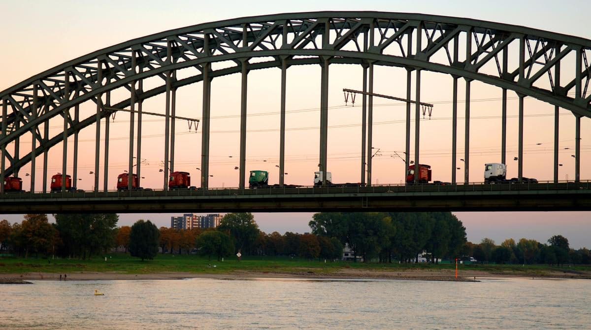 Eine Reihe bunter Lastwagen überquert bei Sonnenuntergang eine große gewölbte Stahlbrücke über einen Fluss, im Hintergrund sind Bäume und Gebäude zu sehen.