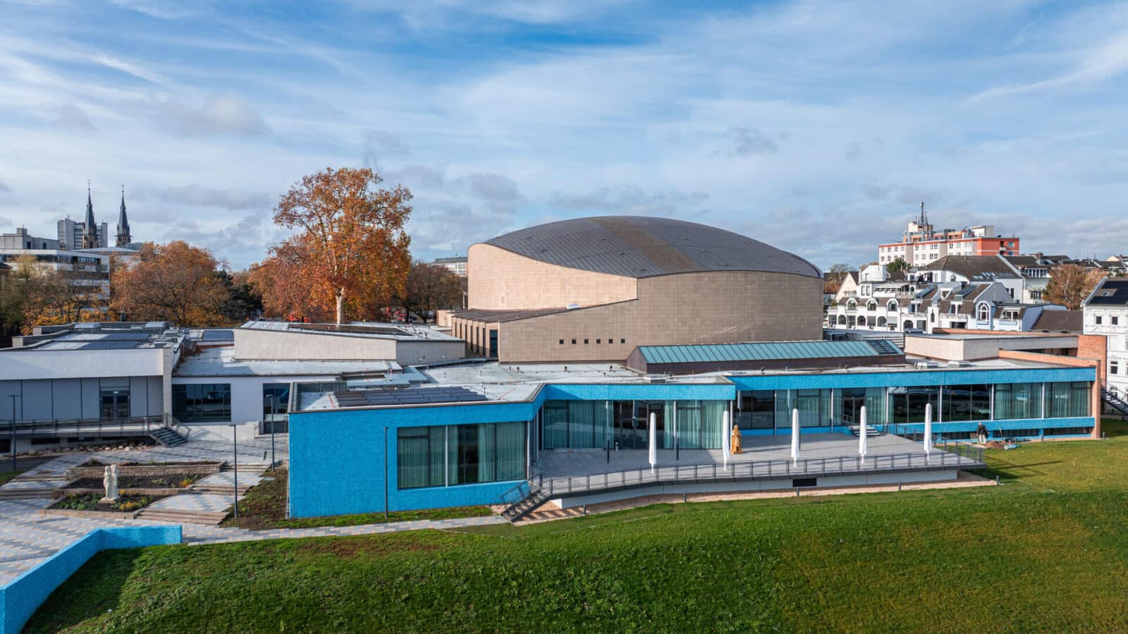 Moderner Gebäudekomplex mit einem großen, runden, beigen Auditorium und einem hellblauen, rechteckigen Gebäude davor, auf einer Rasenfläche mit Bäumen und anderen städtischen Gebäuden im Hintergrund unter einem teilweise bewölkten Himmel.