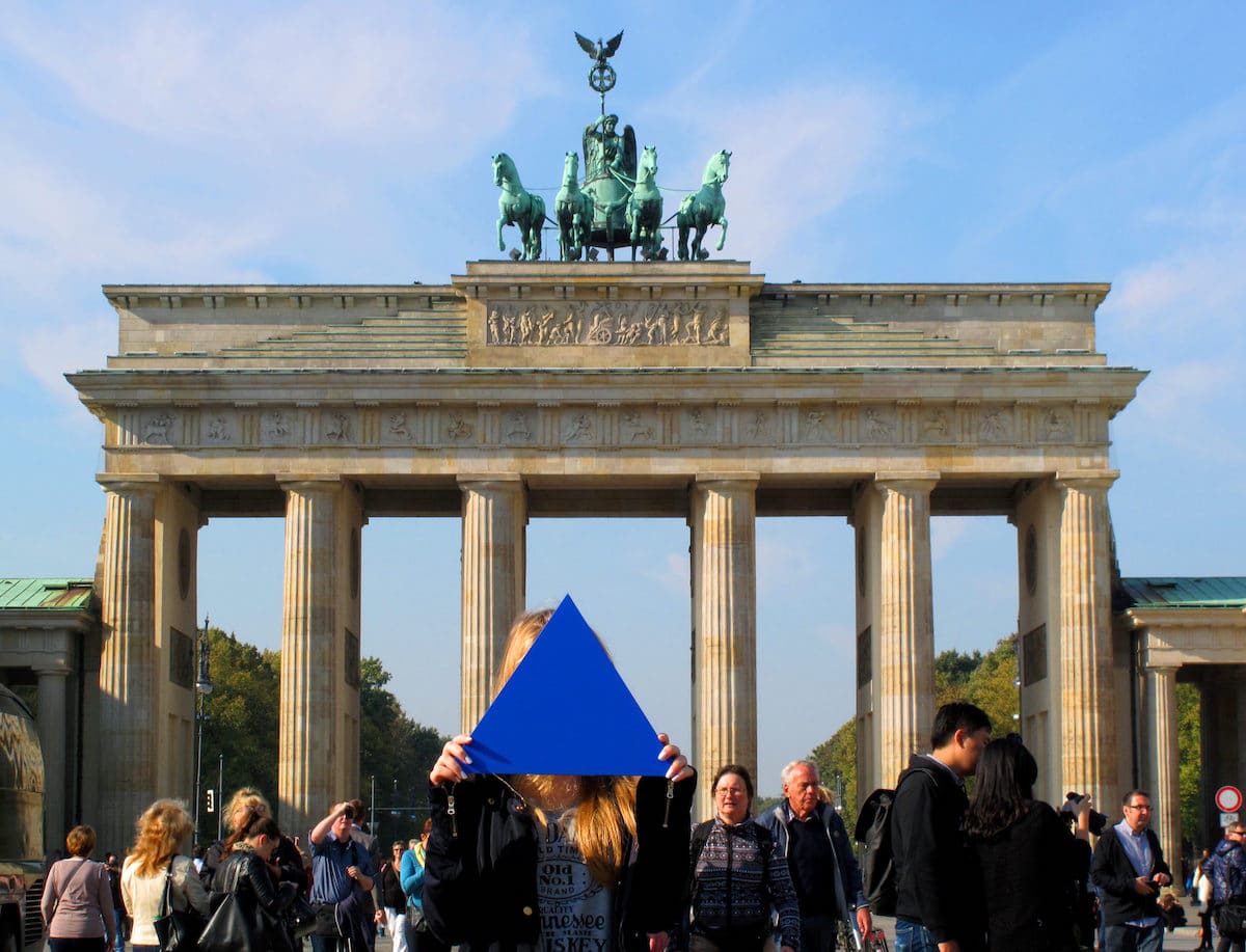 Eine Person hält sich ein großes blaues Dreieck vor das Gesicht und steht an einem sonnigen Tag in einer Menschenmenge vor dem Brandenburger Tor in Berlin.