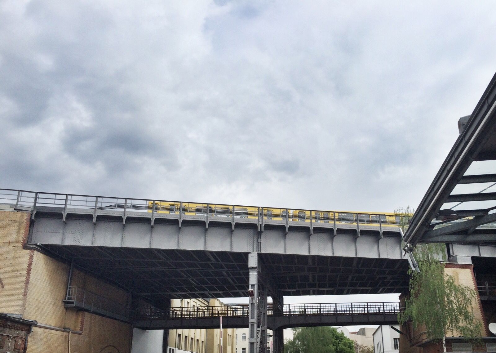 Ein gelber Zug fährt unter einem wolkenverhangenen Himmel über eine erhöhte Stahlbrücke, unter der und seitlich Gebäude und Bäume zu sehen sind.
