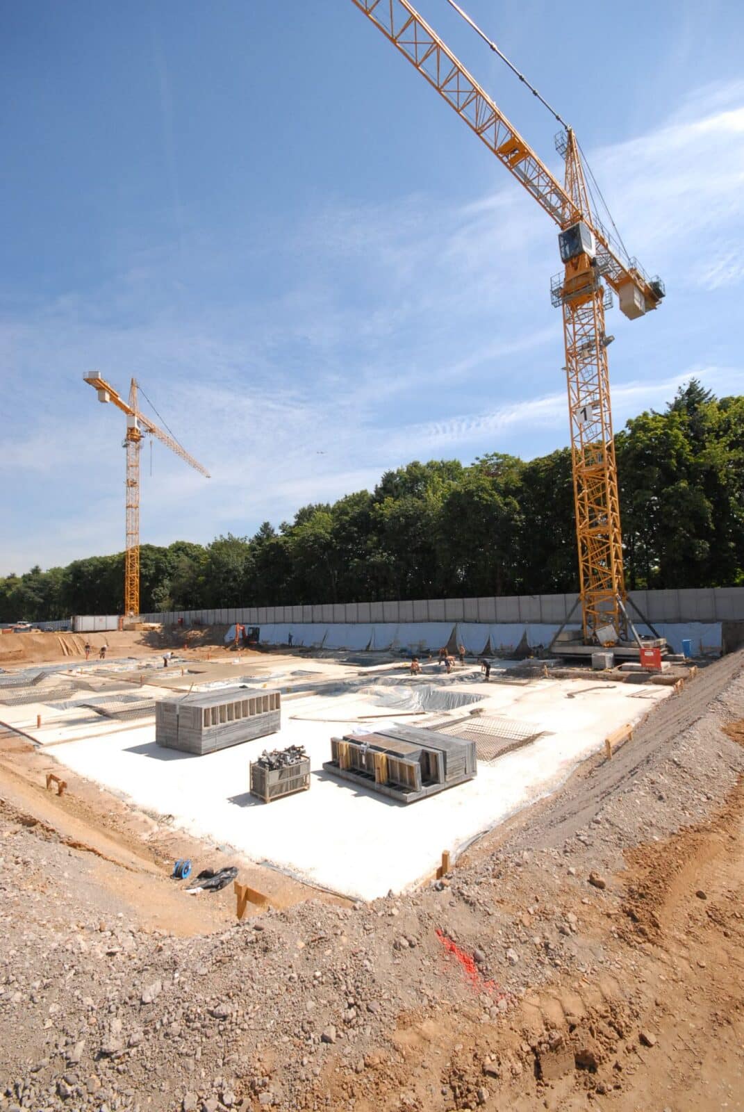 Eine Baustelle mit zwei großen gelben Turmdrehkränen, laufenden Betonfundamenten, Stapeln von Baumaterialien und einer Kulisse aus grünen Bäumen unter einem blauen Himmel.