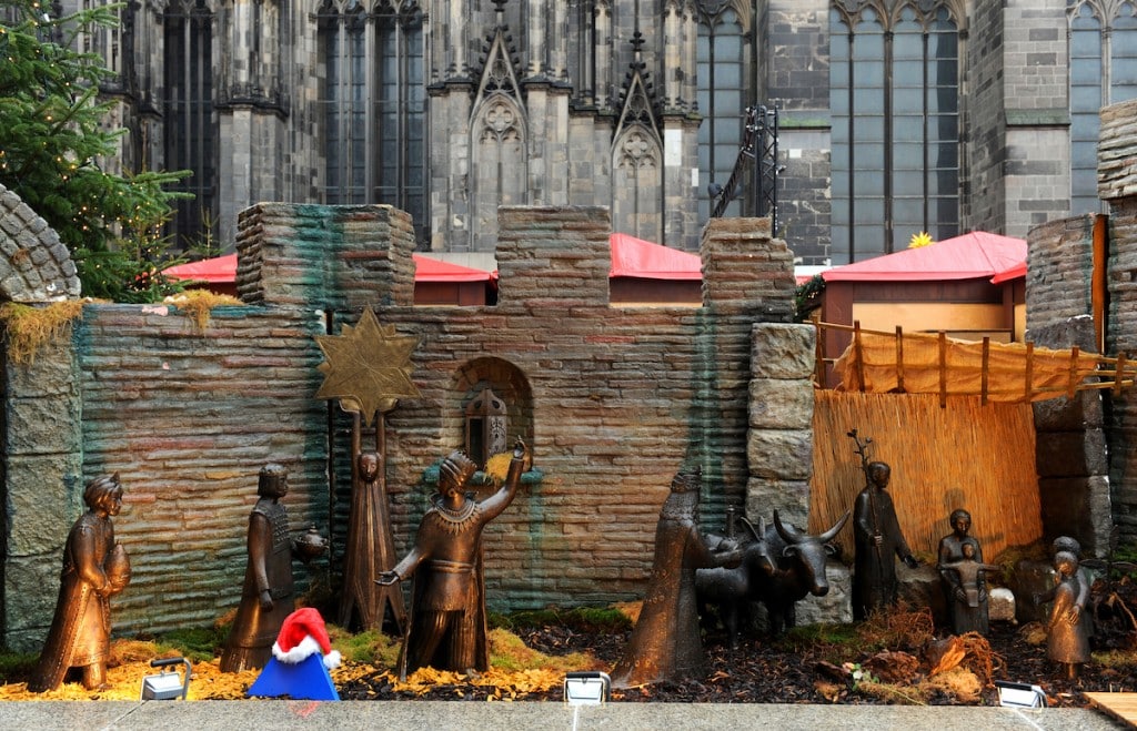Eine Krippe mit Bronzefiguren, Tieren und einem Stern ist vor einer Steinmauer aufgestellt. Auf der linken Seite steht eine Figur mit einer kleinen roten Weihnachtsmannmütze. Im Hintergrund ist die Architektur der Kathedrale zu sehen.