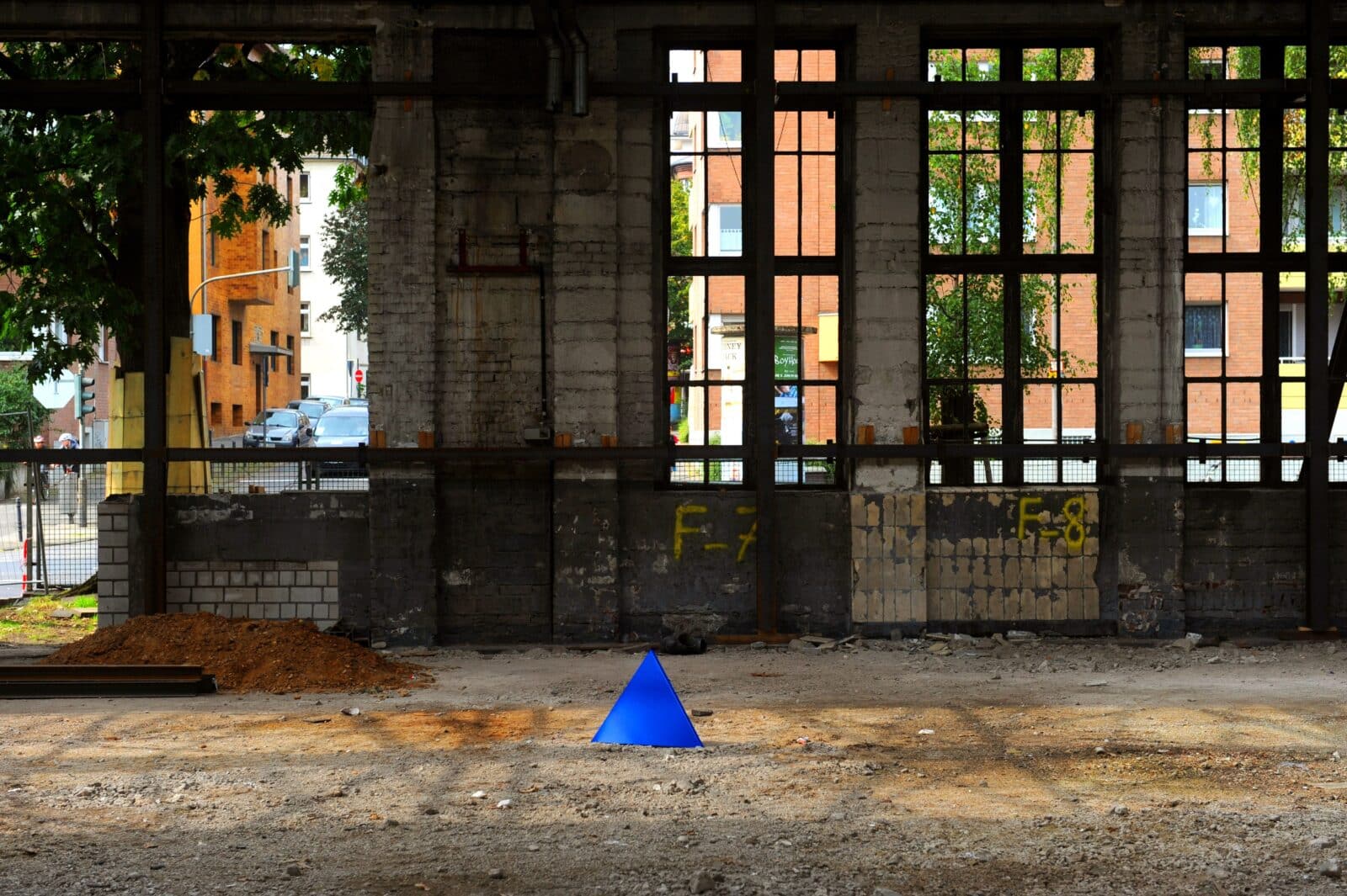 Ein leuchtend blauer Kegel steht auf dem staubigen Boden eines verlassenen Industriegebäudes mit großen Fenstern und freiliegenden Ziegeln, die das Licht hereinlassen und den Blick auf städtische Gebäude und Bäume im Freien freigeben.