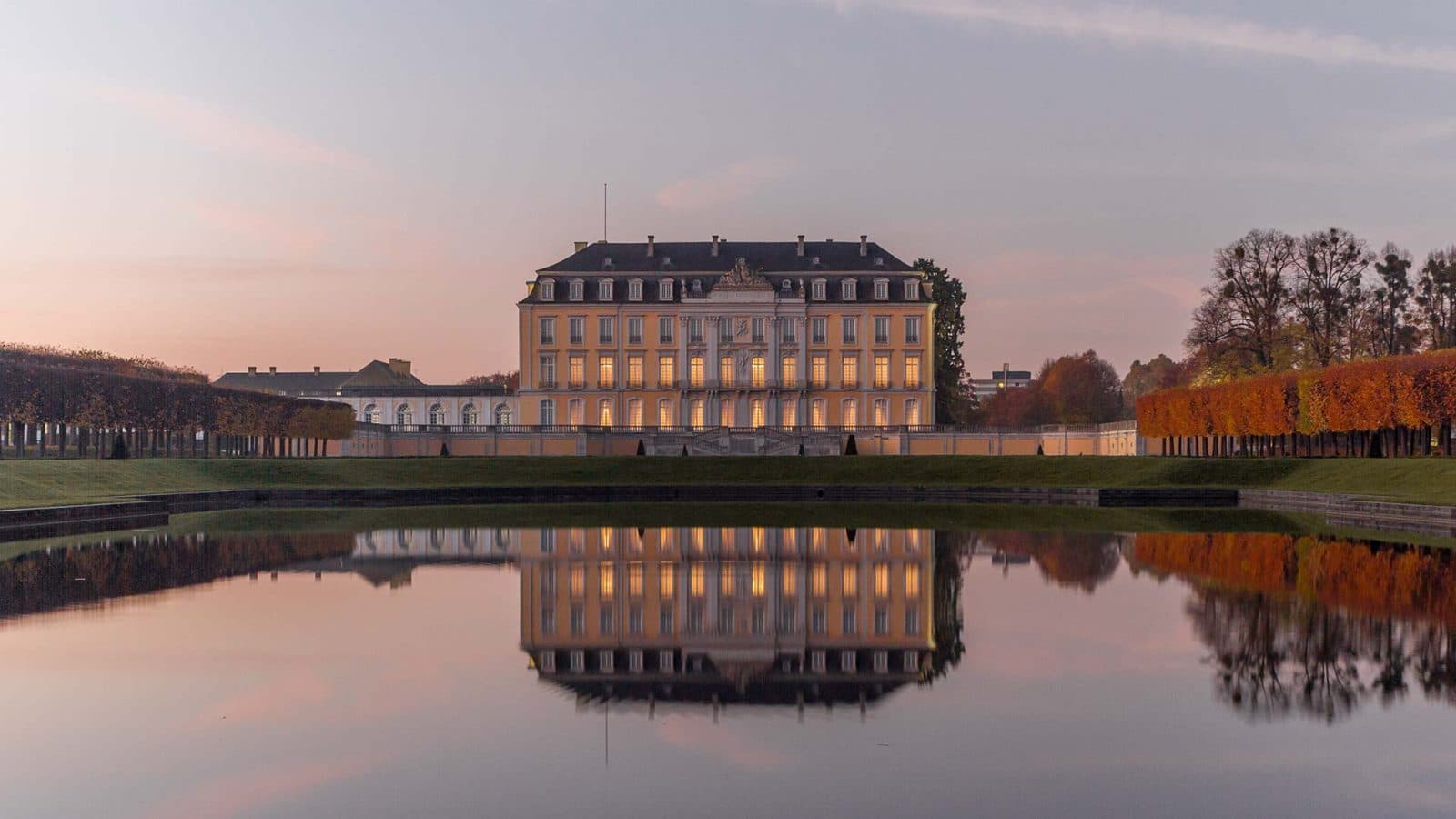 Ein großer, eleganter Palast mit beleuchteten Fenstern spiegelt sich bei Sonnenuntergang in einem ruhigen Teich, eingerahmt von gepflegten Rasenflächen und herbstlich gefärbten Baumreihen auf beiden Seiten.