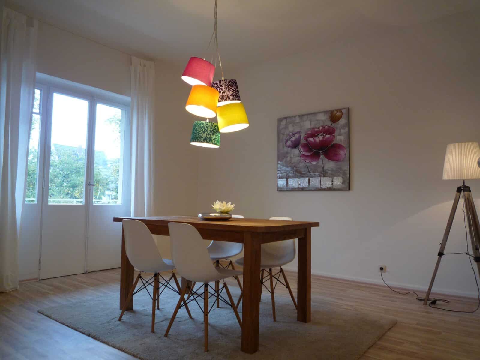 Ein minimalistisches Esszimmer mit einem Holztisch, vier weißen Stühlen, einem beigen Teppich und bunten Pendelleuchten. Ein großes Fenster, eine Stehlampe und florale Wandkunst schmücken den Raum.
