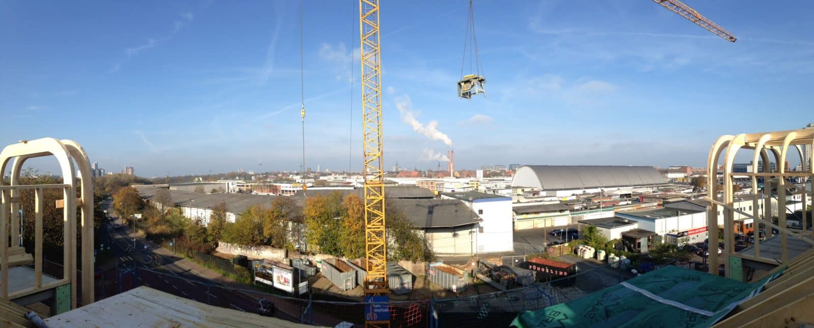 Panoramablick auf ein Industriegebiet mit Lagerhäusern, einem großen Gebäude mit geschwungenem Dach, Baukränen und in der Ferne aufsteigendem Rauch unter blauem Himmel. Einige Bäume säumen die Straßen in der unteren linken Ecke.
