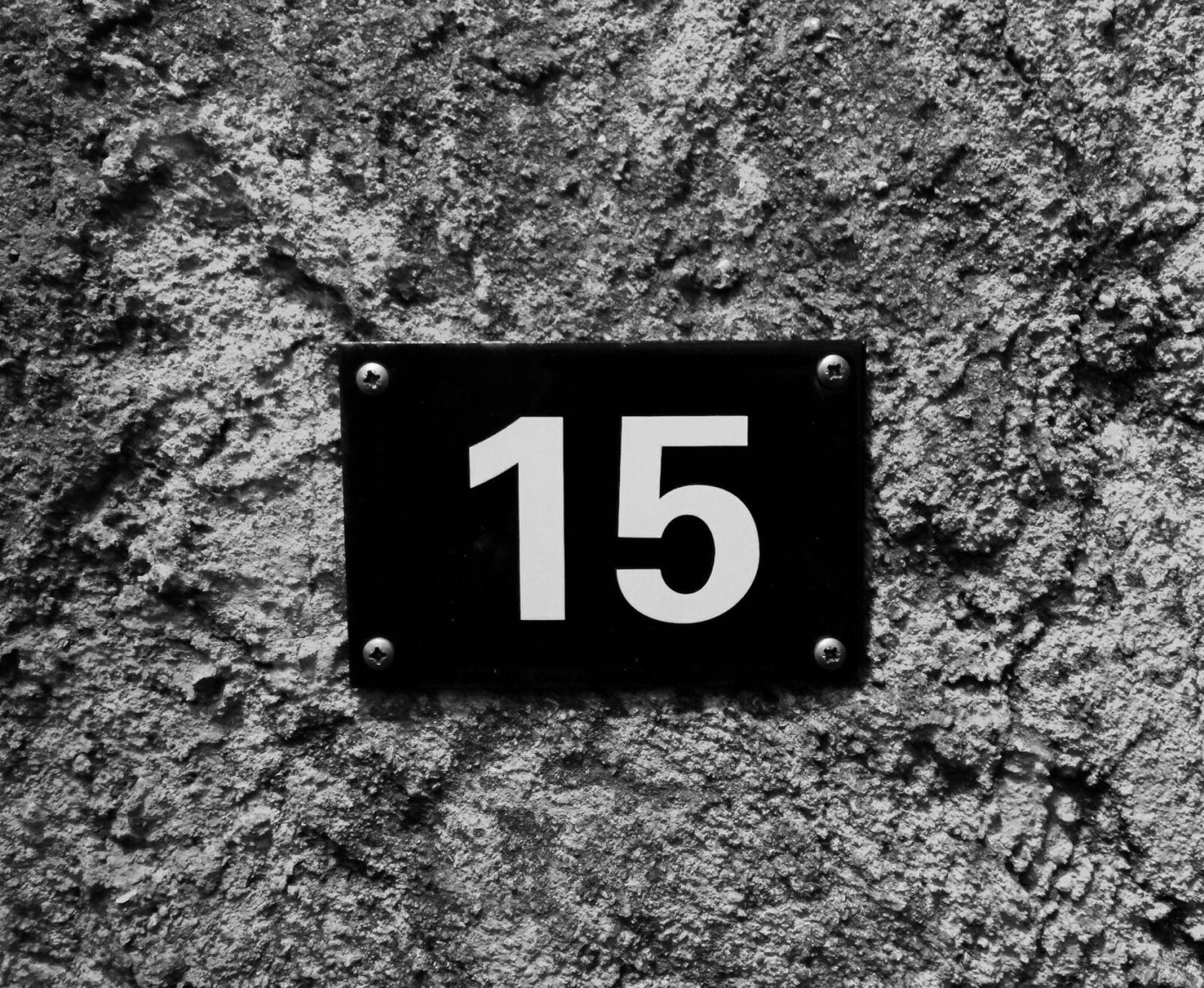Ein schwarzes rechteckiges Schild mit der Zahl 15 in weißer Schrift, das mit vier sichtbaren Schrauben an einer rauen, strukturierten grauen Betonwand befestigt ist.