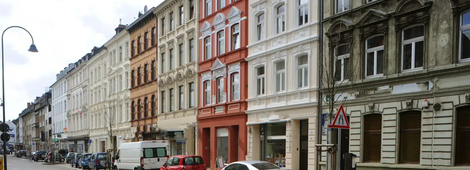 Eine Reihe von mehrstöckigen historischen Gebäuden mit verzierten Fassaden säumen eine Straße. Entlang des Bordsteins sind geparkte Autos zu sehen, und auf dem Bürgersteig sind eine Straßenlaterne und Verkehrsschilder zu erkennen.