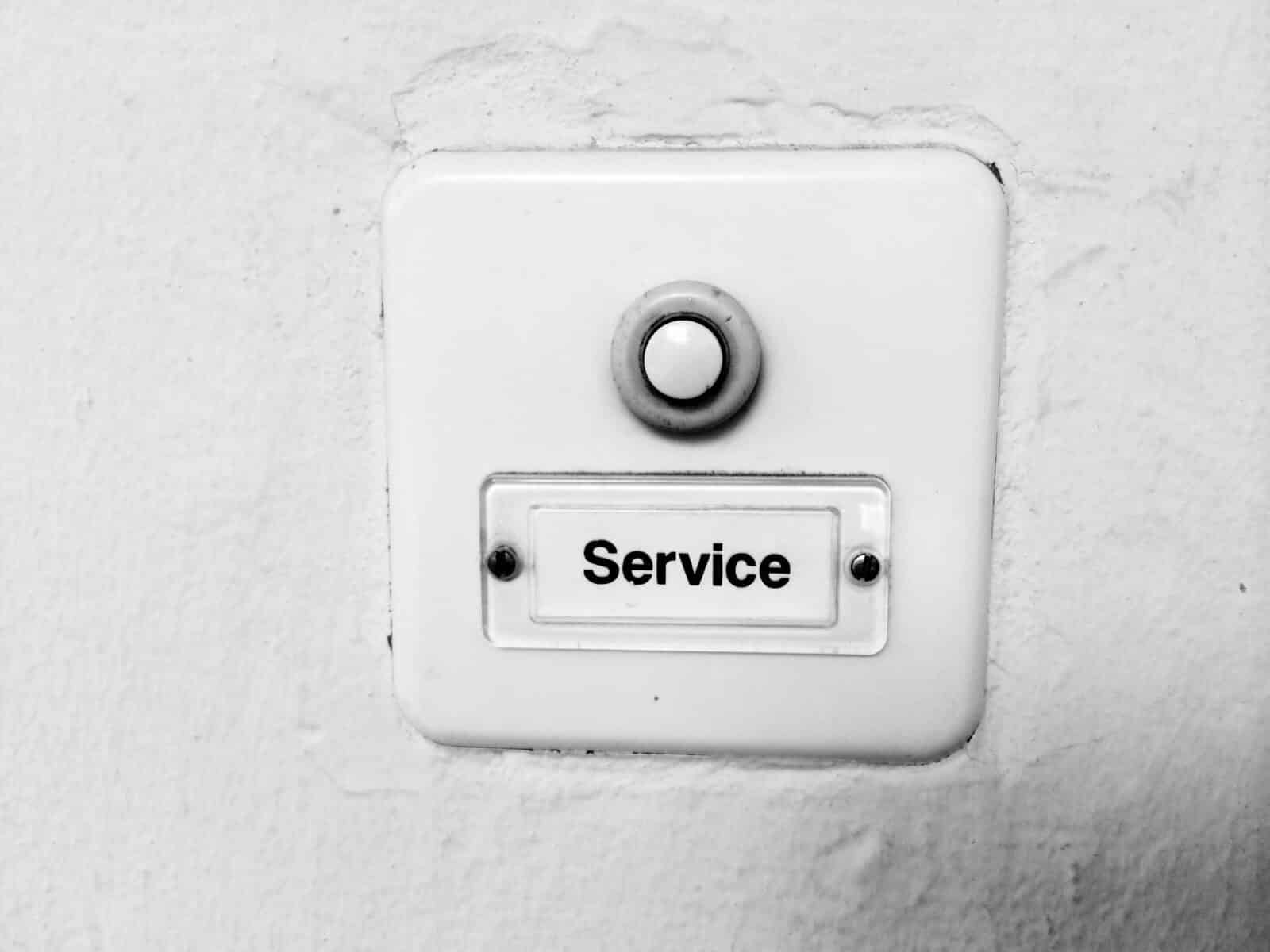 An einer Wand ist ein weißer Klingelknopf angebracht, unter dem ein Schild mit der Aufschrift "Service" in schwarzen Buchstaben angebracht ist. Die Wand weist einige Gebrauchsspuren auf.