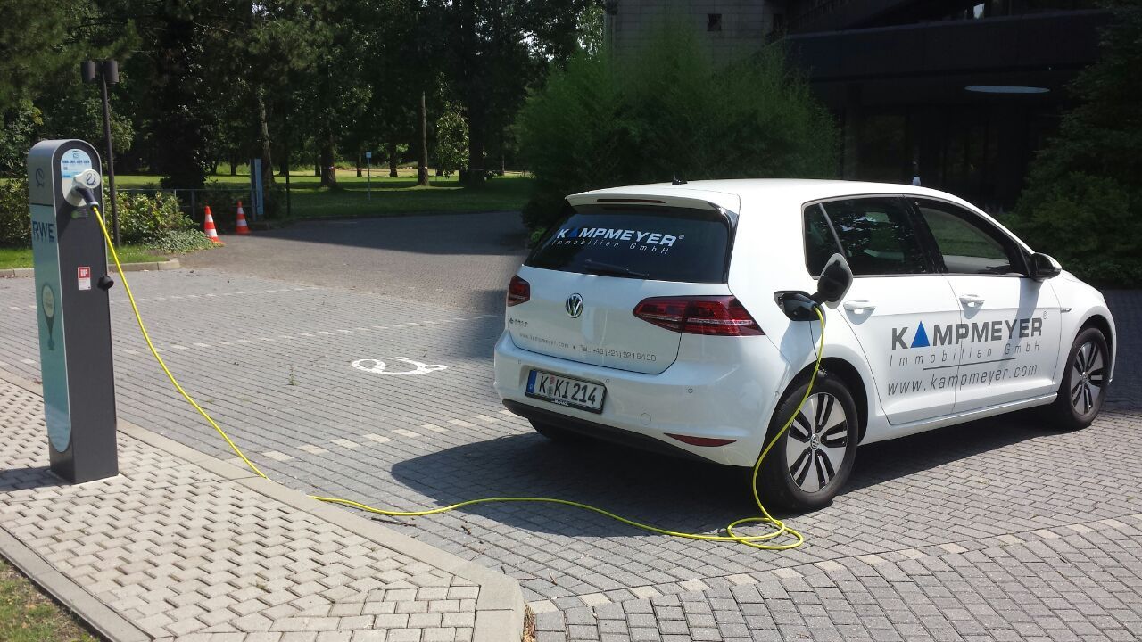 Ein weißes Elektroauto mit KAMPMEYER-Schriftzug parkt und lädt an einer Ladestation für Elektrofahrzeuge auf einem gepflasterten, begrünten Parkplatz.