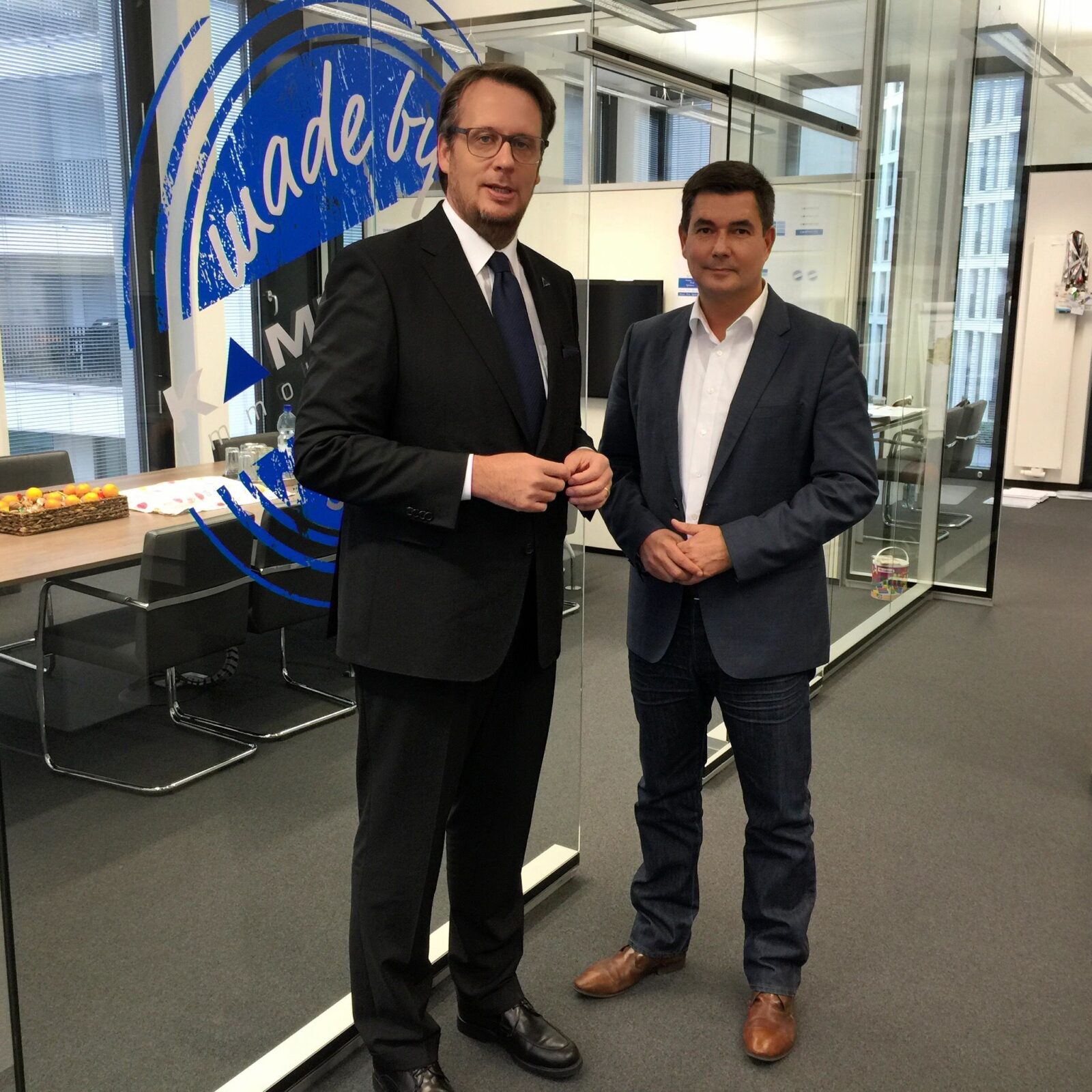 Zwei Männer stehen in einem modernen Büro mit Glaswänden. Der eine trägt Anzug und Krawatte, der andere einen dunklen Blazer mit einem weißen Hemd und Jeans. Hinter ihnen sind ein Konferenztisch und ein großes blaues Logo an der Glaswand zu sehen.