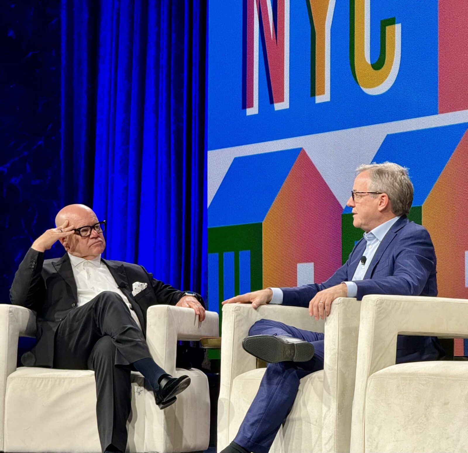 Zwei Männer in Anzügen sitzen auf weißen Stühlen auf einer Bühne und führen ein Gespräch. Hinter ihnen befindet sich ein farbenfroher Hintergrund mit großen Buchstaben, auf denen "NYC" steht, und stilisierten Formen von Gebäuden.