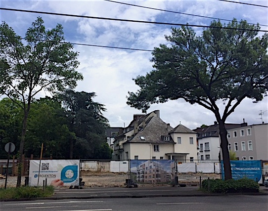 Eine eingezäunte Baustelle mit Bannern vor einem weißen Haus, umgeben von Bäumen und anderen Wohngebäuden unter einem teilweise bewölkten Himmel.