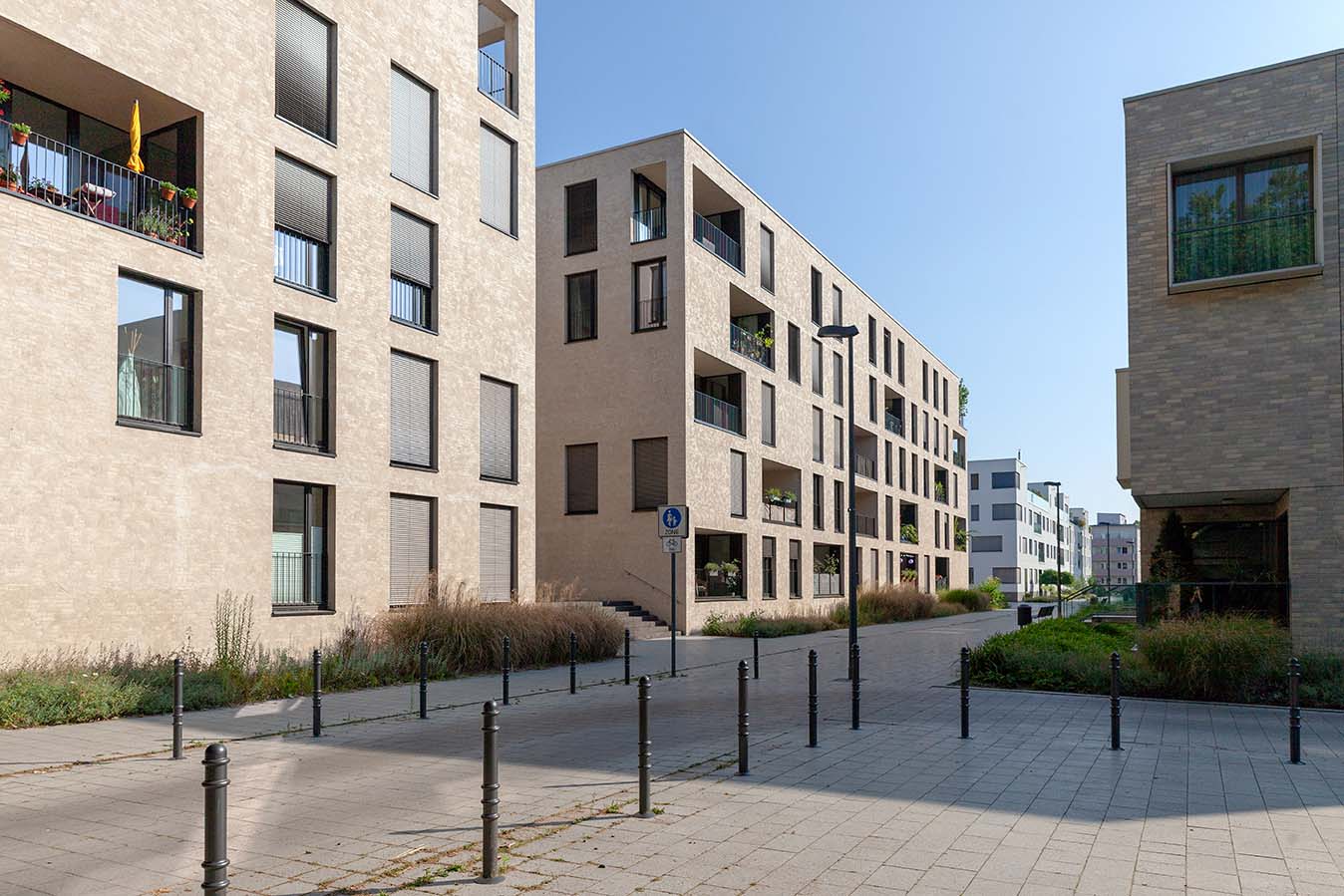 Eine moderne Wohnstraße mit beigefarbenen Backsteinwohnungen, großen Fenstern, Balkonen und Grünanlagen unter einem strahlend blauen Himmel. Schwarze Poller säumen den breiten gepflasterten Gehweg.