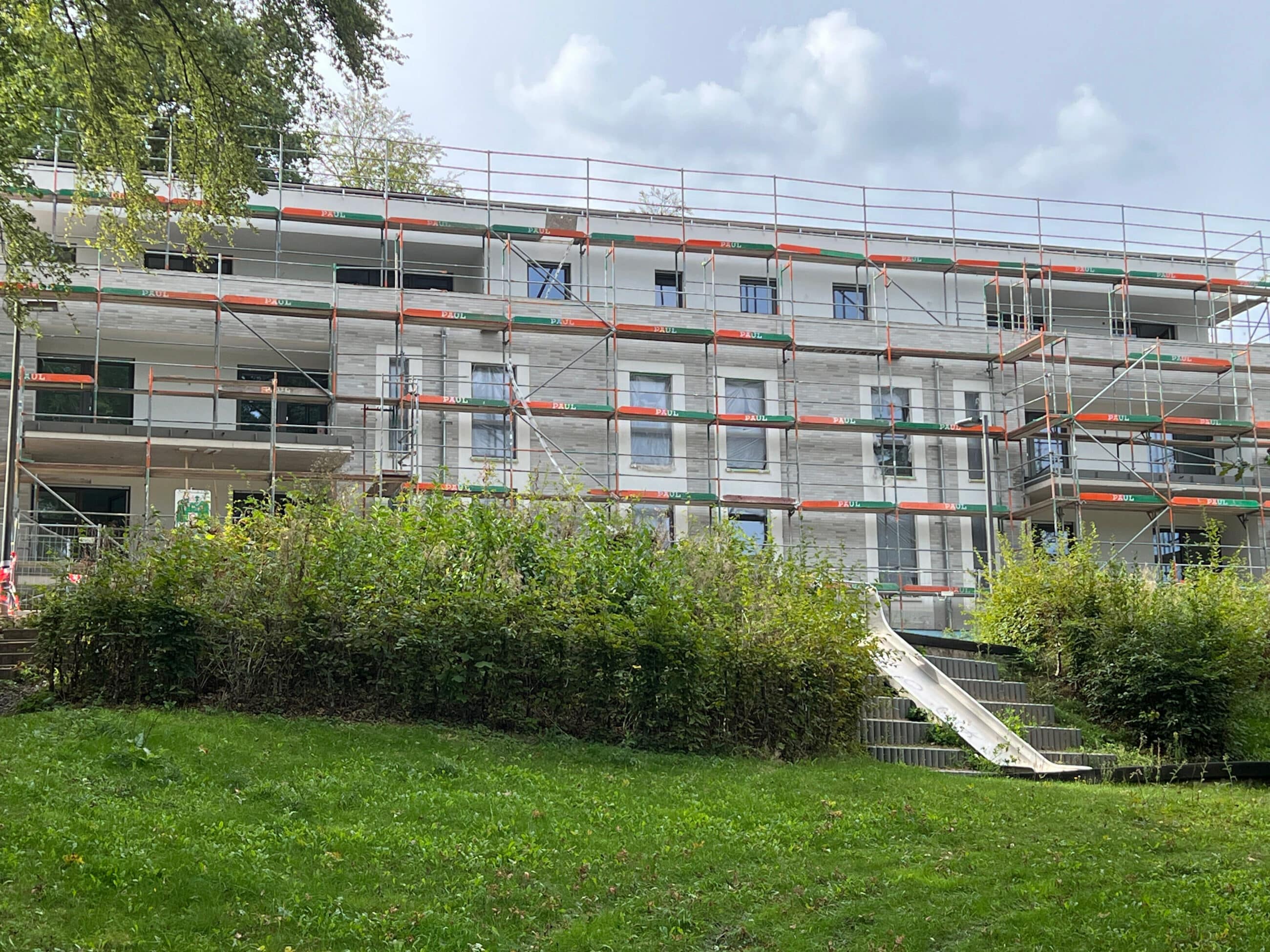 Ein im Bau befindliches dreistöckiges Gebäude ist von Gerüsten umgeben. Im Vordergrund sind grüne Büsche und Gras zu sehen, während darüber ein bewölkter Himmel zu erkennen ist.