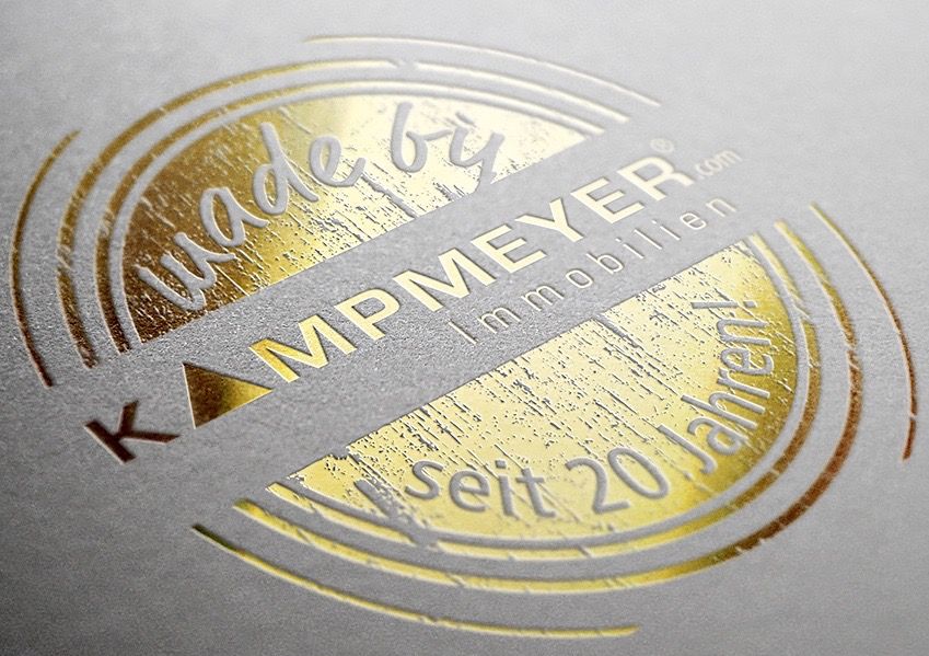Auf einer strukturierten hellgrauen Oberfläche ist ein kreisförmiges Logo in Goldprägung mit der Aufschrift made by KAMPMEYER Immobilien seit 20 Jahren! angebracht. Der Text und das Design haben eine glänzende, reflektierende Oberfläche.