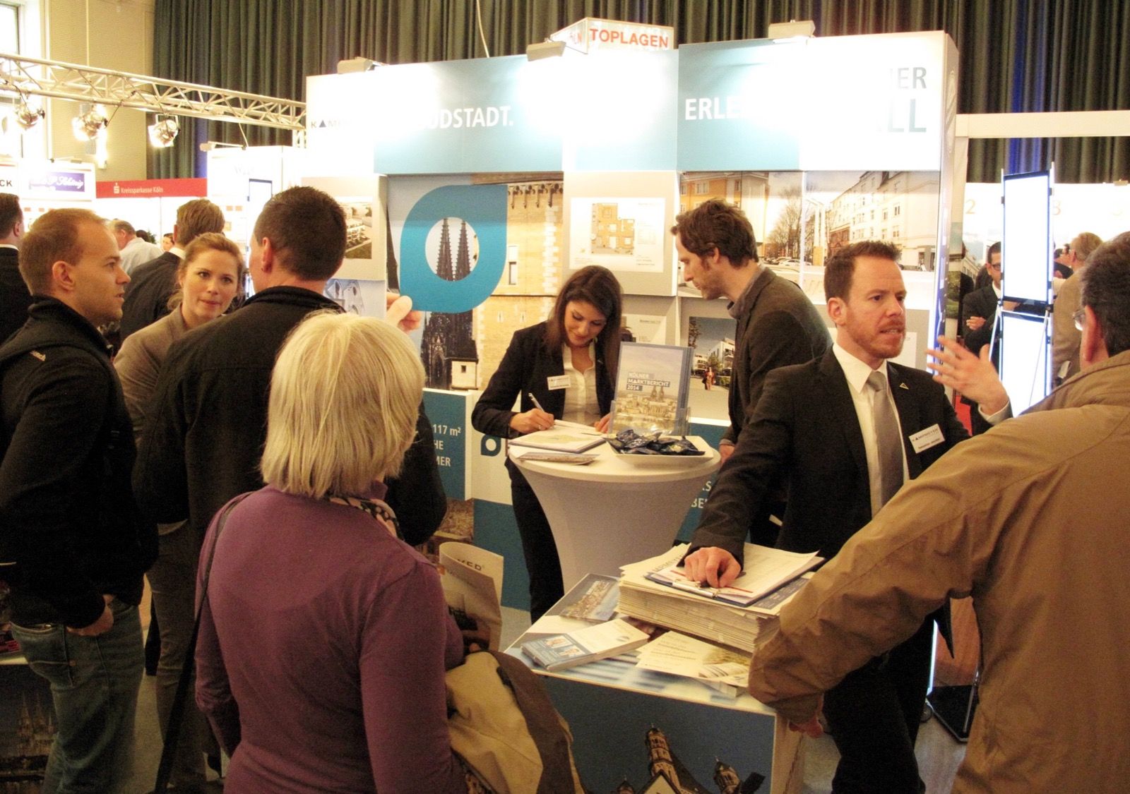 Eine Gruppe von Menschen versammelt sich um einen Stand auf einer belebten Messe. Einige Personen sprechen mit Ausstellern, während andere sich Broschüren und Displays ansehen. Ein Mann im Anzug spricht und gestikuliert am Empfangstresen.