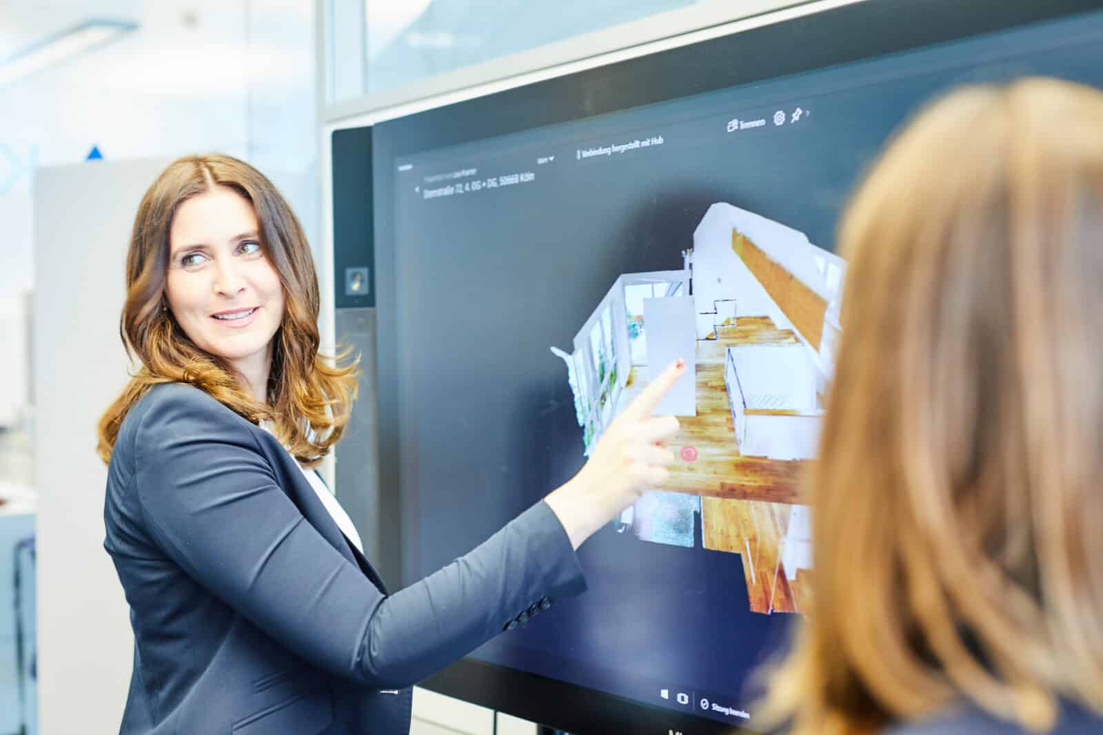 Eine Frau in einem Blazer zeigt auf einen großen Bildschirm, auf dem ein 3D-Grundriss zu sehen ist, während eine weitere Person daneben steht. Die beiden scheinen in einer modernen Büroumgebung über das digitale Modell zu diskutieren oder es zu präsentieren – vielleicht arbeiten sie gemeinsam mit einem Immobilienmakler aus der Kölner Innenstadt an Gestaltungsideen für eine Immobilie.