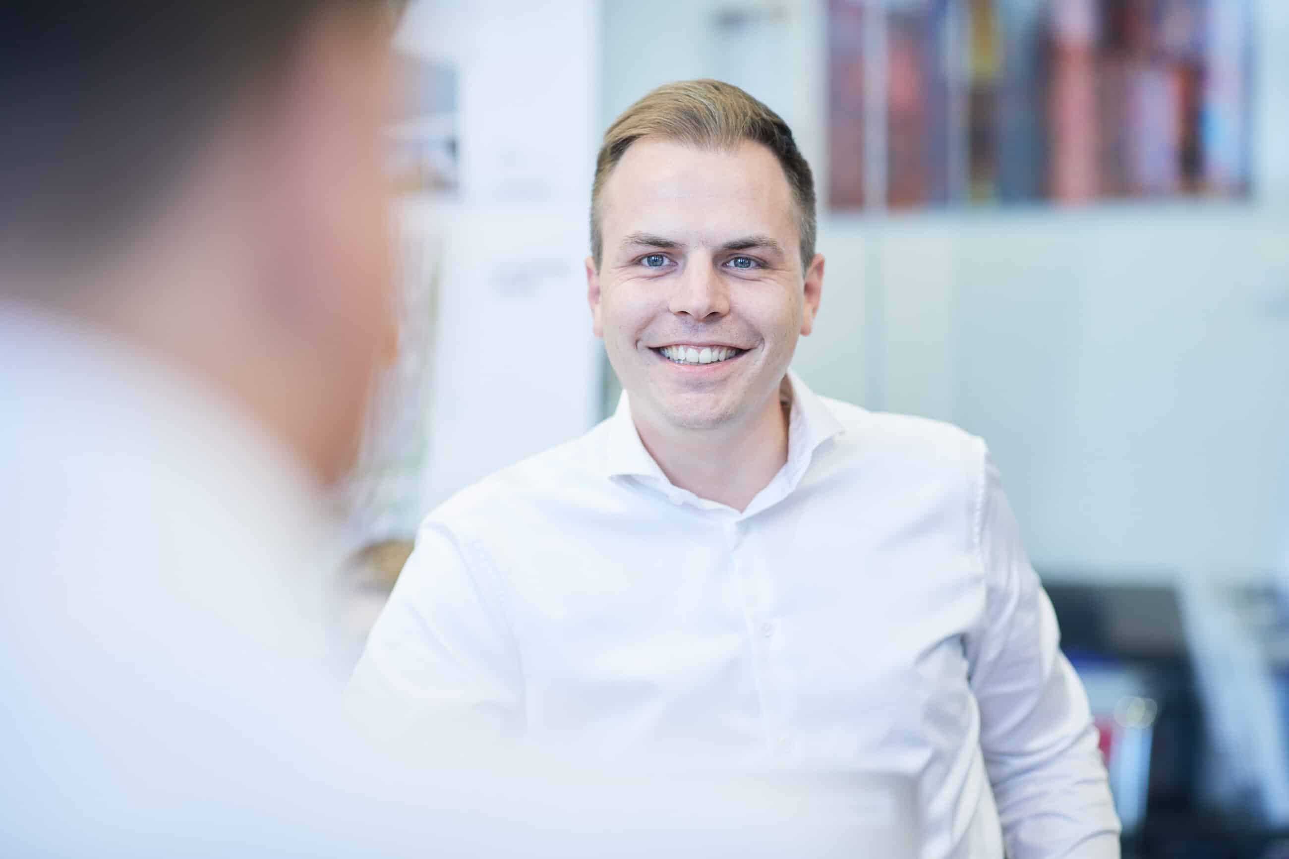 Ein Mann in einem weißen Hemd lächelt, während er jemanden in einer Büroumgebung ansieht. Der Hintergrund ist unscharf und hebt seinen freundlichen Ausdruck hervor.