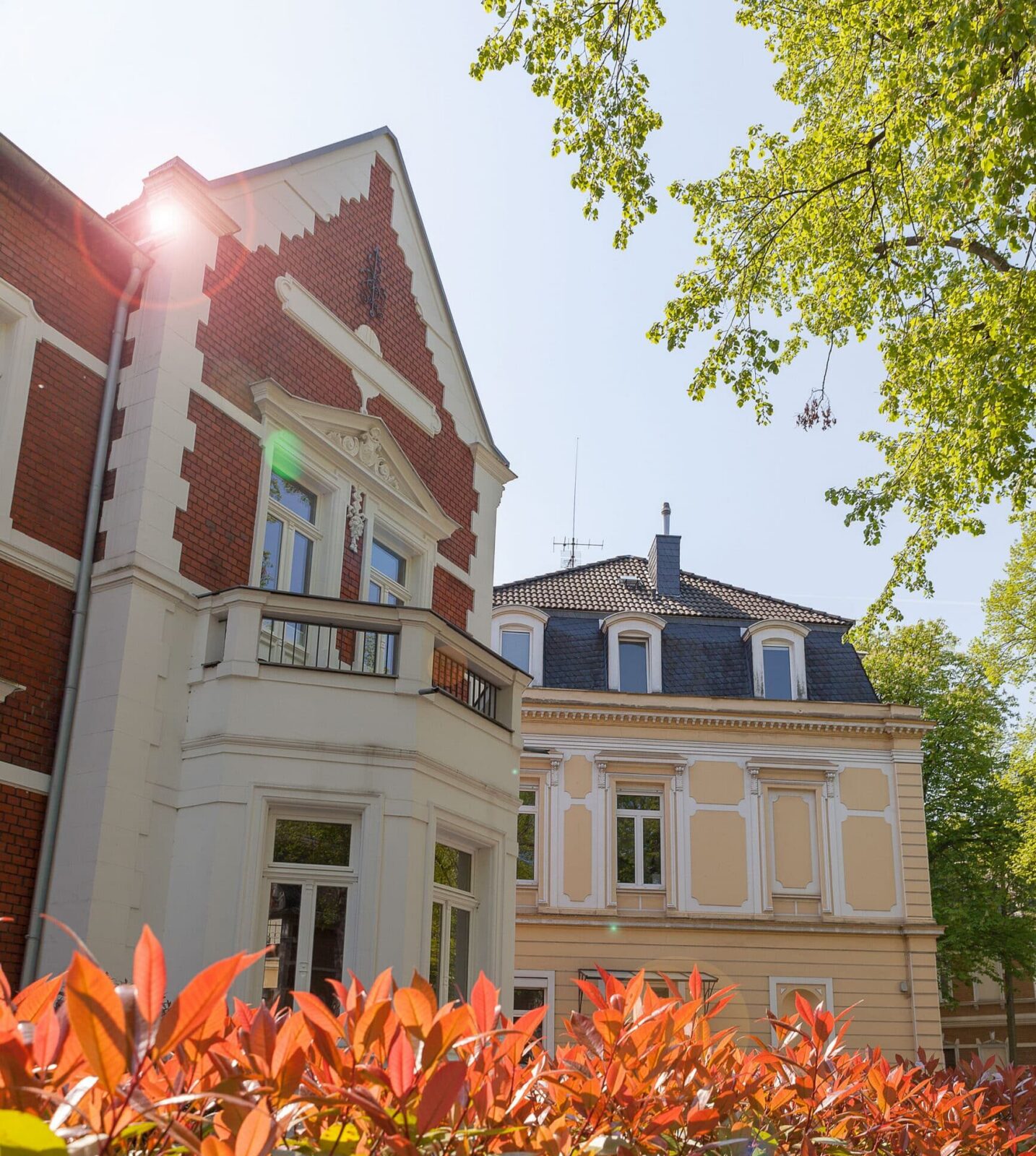 Zwei elegante, historische Gebäude mit verzierten Fassaden stehen an einem sonnigen Tag nebeneinander. Rotblättrige Sträucher und grüne Bäume rahmen die Szene ein, und das Sonnenlicht erzeugt in der Nähe des Dachs des roten Backsteingebäudes einen Streiflicht-Effekt.