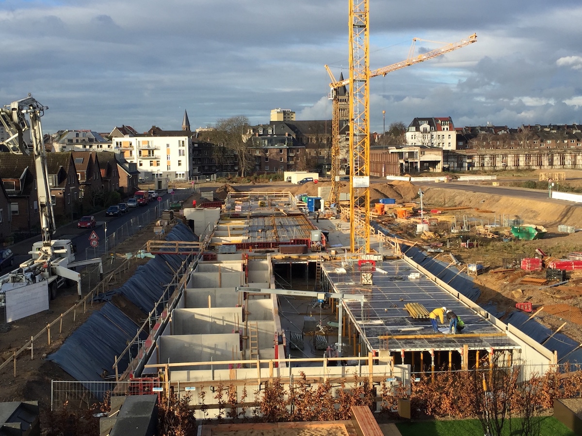 Eine Baustelle mit Betonstrukturen, Gerüsten und einem hohen gelben Kran; im Hintergrund sind Gebäude und Häuser unter einem teilweise bewölkten Himmel zu sehen.