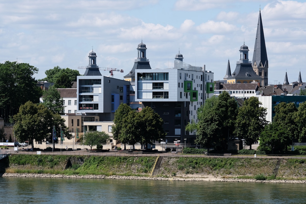 Moderne und historische Gebäude, darunter eine Kirche mit einem hohen Kirchturm, säumen das Flussufer unter einem teilweise bewölkten Himmel, mit Bäumen und Grünpflanzen im Vordergrund.