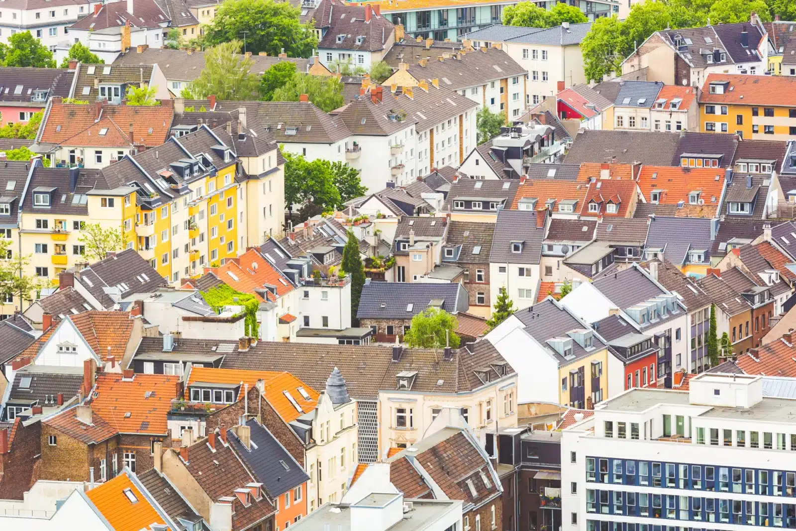 Luftaufnahme eines dichten Stadtviertels mit einer Mischung aus bunten Wohngebäuden, meist mit roten und grauen Ziegeldächern, umgeben von Bäumen und Grünanlagen.