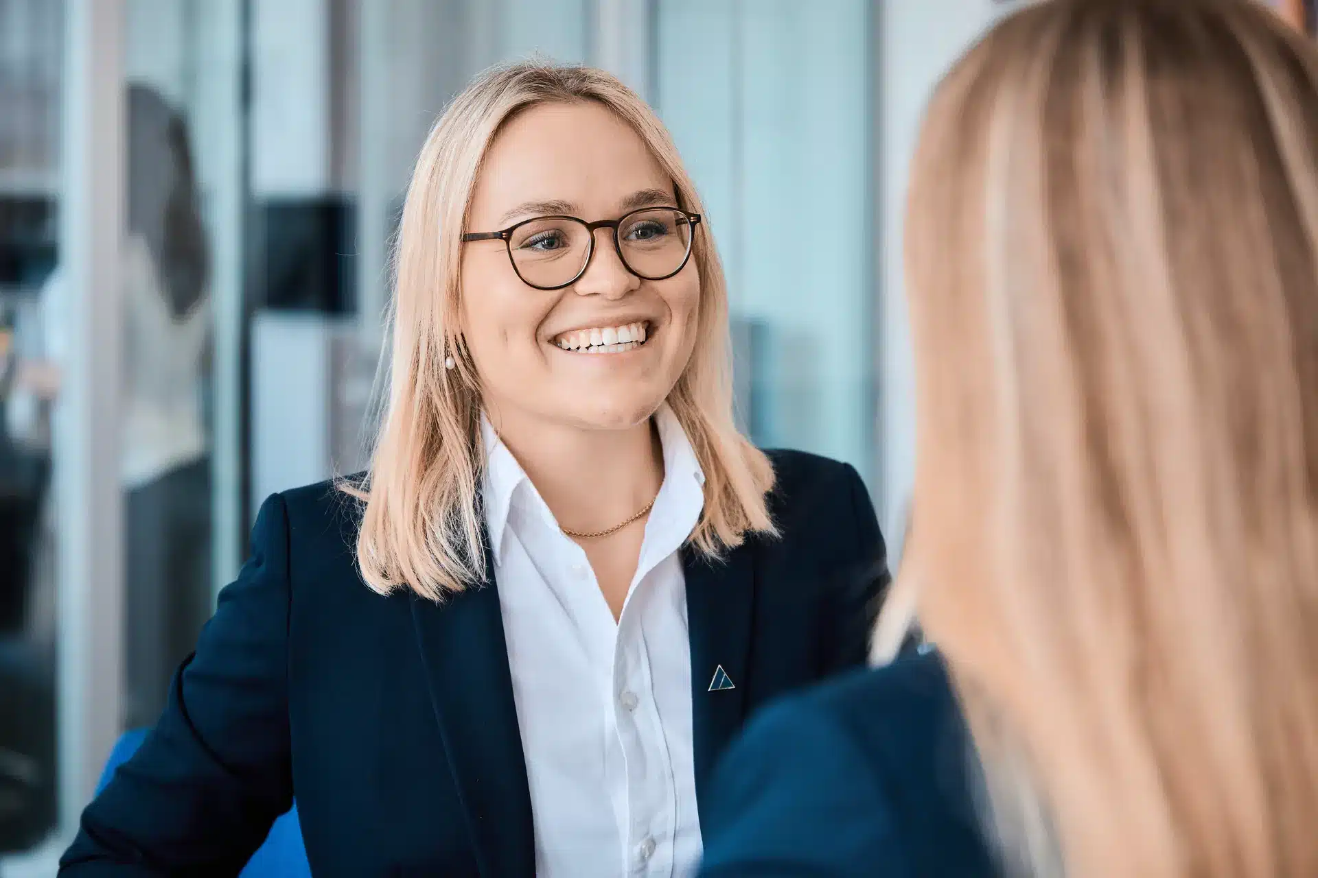Eine lächelnde Frau mit blondem Haar, die eine Brille, ein weißes Hemd und einen dunklen Blazer trägt, sitzt in einem Büro und spricht mit einer anderen Person.