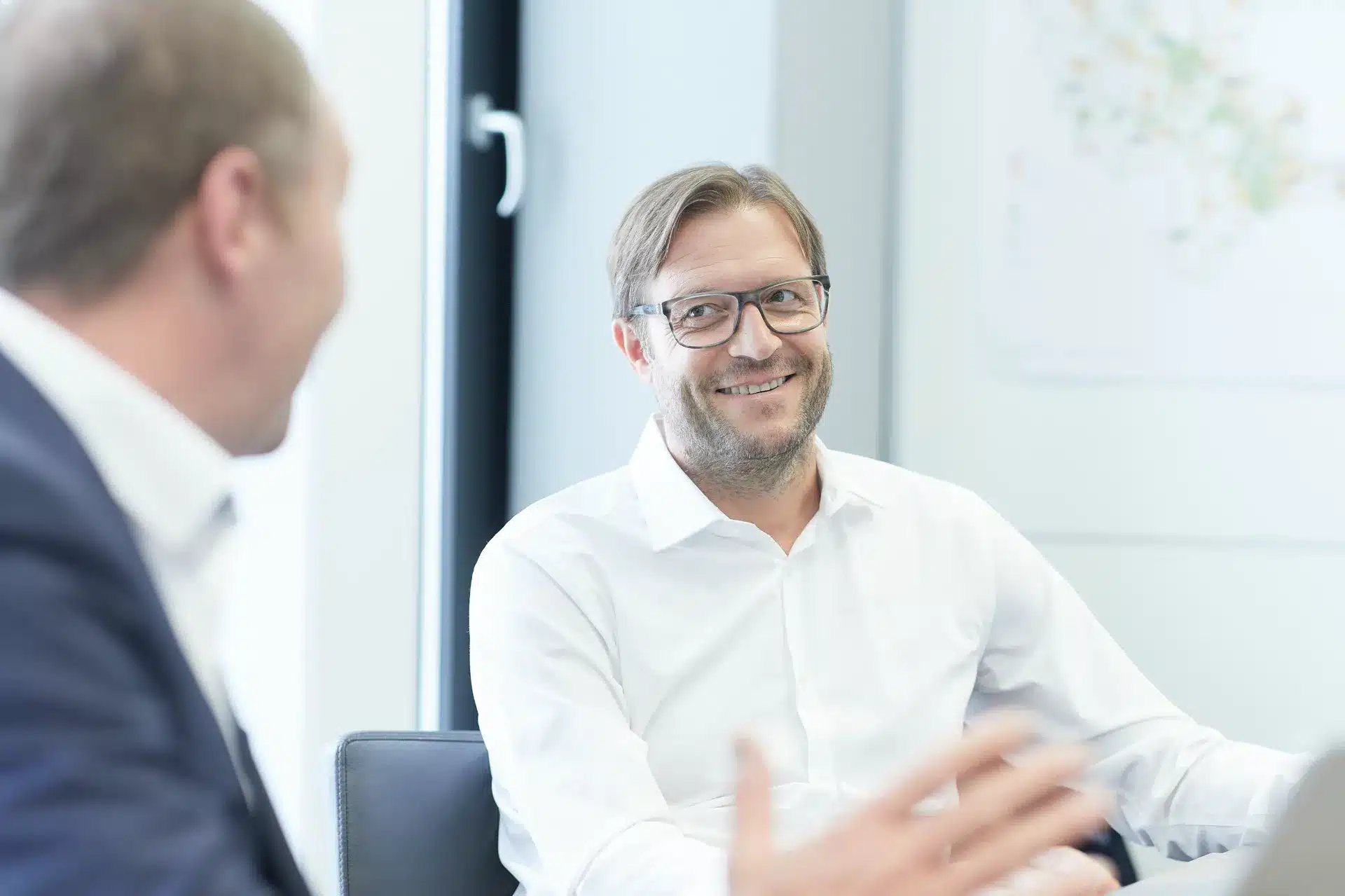 Zwei Männer in Geschäftskleidung führen ein Gespräch in einem Büro. Der eine Mann, der eine Brille und ein weißes Hemd trägt, lächelt und blickt den anderen Mann an, der sich leicht verschwommen im Vordergrund befindet.