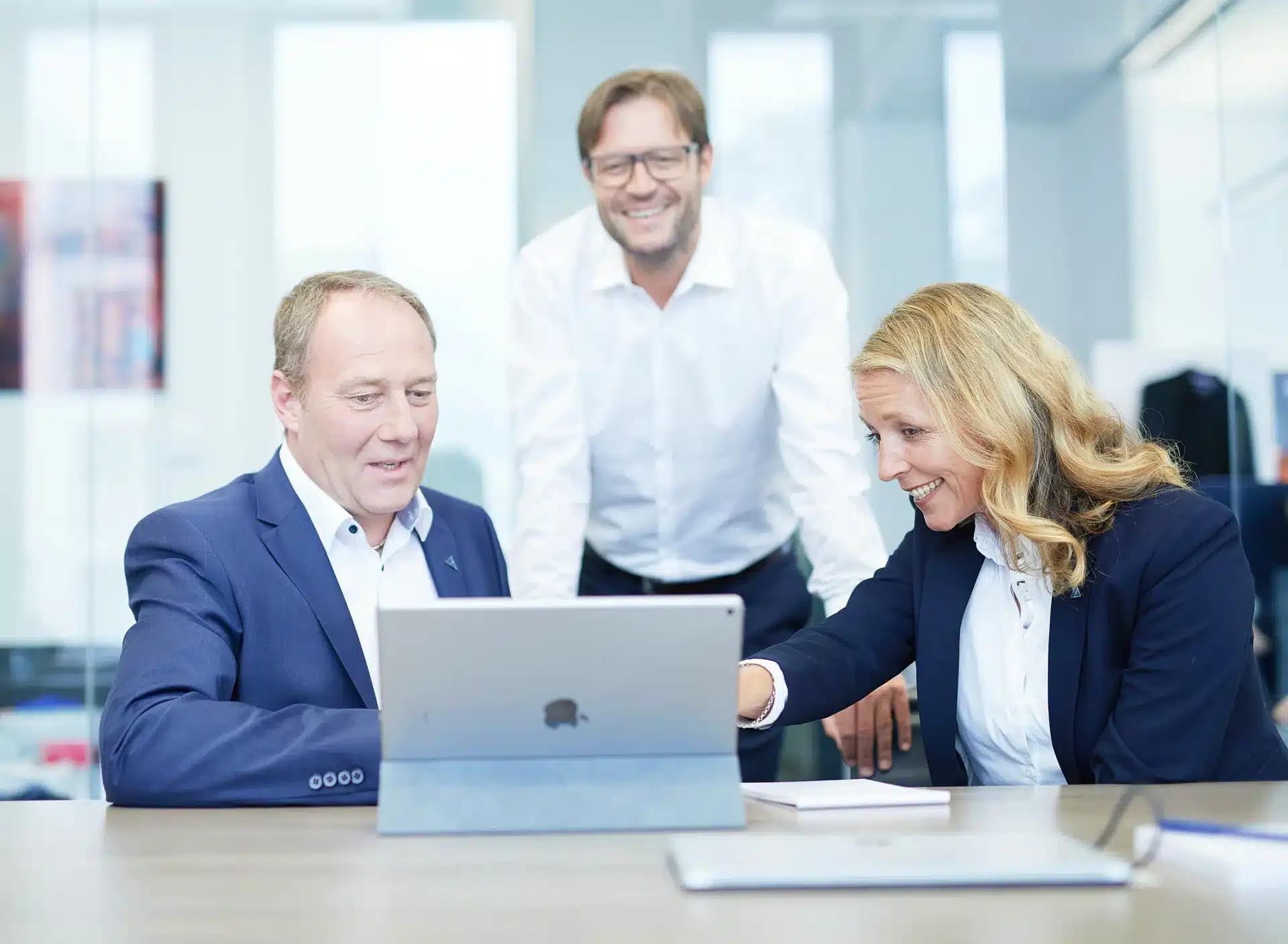 Drei Geschäftsleute, zwei Männer und eine Frau, sitzen an einem Tisch in einem modernen Büro, lächeln und schauen gemeinsam auf einen Laptop-Bildschirm. Die Atmosphäre scheint kollegial und positiv zu sein.