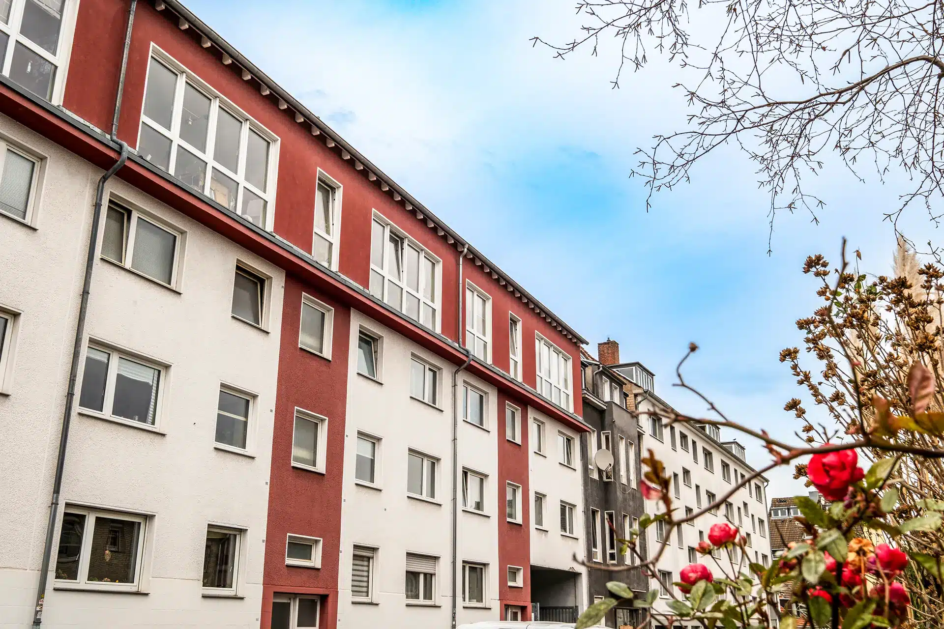 Blick aus geringer Höhe auf ein Mehrfamilienhaus mit weißen und roten Außenwänden, mehreren Fenstern und blühenden roten Blumen im Vordergrund unter einem teilweise bewölkten Himmel.