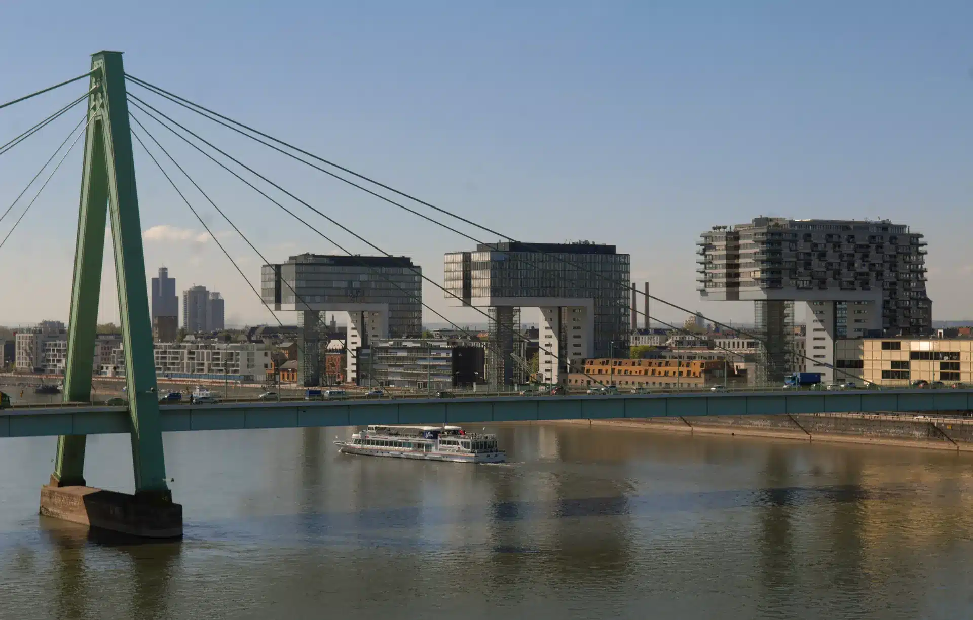 Eine moderne Schrägseilbrücke überspannt einen Fluss, unter der ein Boot hindurchfährt. Im Hintergrund sind drei kranförmige Gebäude und andere städtische Strukturen unter einem klaren blauen Himmel zu sehen.