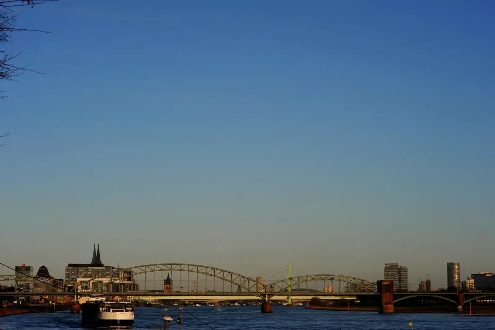 Eine Stadtlandschaft in der Abenddämmerung zeigt eine große Metallbrücke, die einen Fluss überspannt, mit Gebäuden auf beiden Seiten. Im Vordergrund liegt ein Boot vor Anker, und der Himmel ist klar und blau.