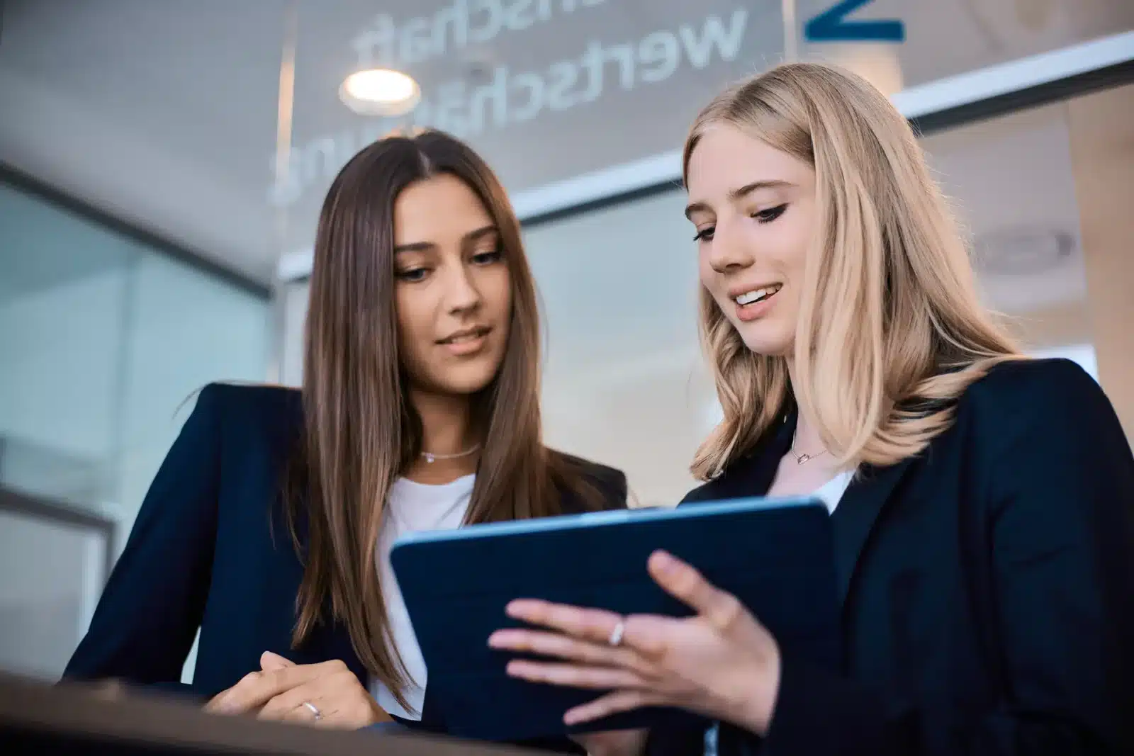 Zwei junge Frauen in Geschäftskleidung lächeln, während sie in einer modernen Büroumgebung gemeinsam auf ein Tablet schauen.