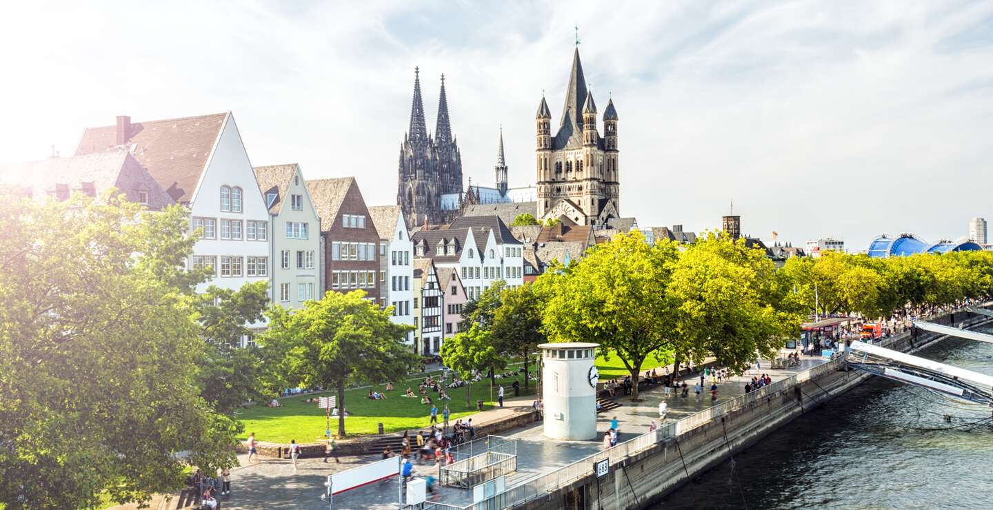 Eine sonnige Uferszene in Köln, Deutschland, mit den Zwillingstürmen des Kölner Doms, der St. Martinskirche, bunten historischen Gebäuden, grünen Bäumen und Menschen, die an der Promenade am Wasser spazieren gehen.