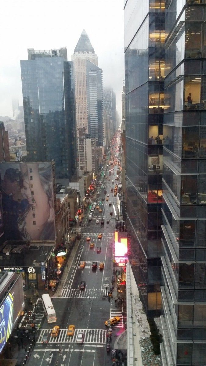 Eine verregnete Straße in New York, gesäumt von hohen Wolkenkratzern und belebt von Autos und gelben Taxis; Fußgänger gehen auf den Bürgersteigen und helle Werbetafeln erhellen die Szene.