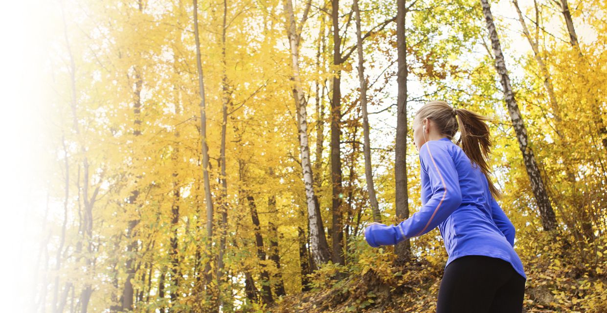 Eine Frau in einem blauen langärmeligen Hemd joggt durch einen Wald mit hohen Bäumen und gelben Herbstblättern, die eine helle und lebendige Herbstatmosphäre schaffen.