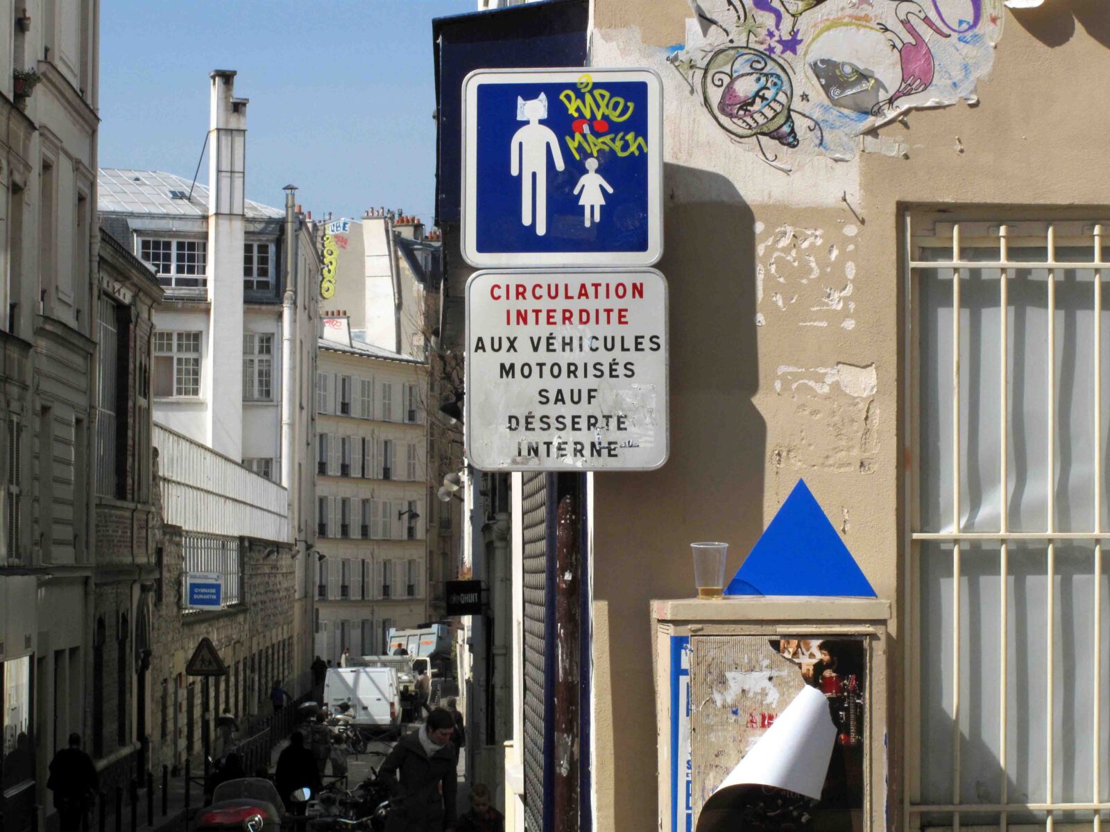Ein französisches Straßenschild mit Aufklebern und Graffiti zeigt ein Fußgängersymbol und die Angabe CIRCULATION INTERDITE AUX VÉHICULES MOTORISÉS SAUF DESSERTE INTERNE auf einer engen Straße mit Gebäuden und Menschen im Hintergrund.