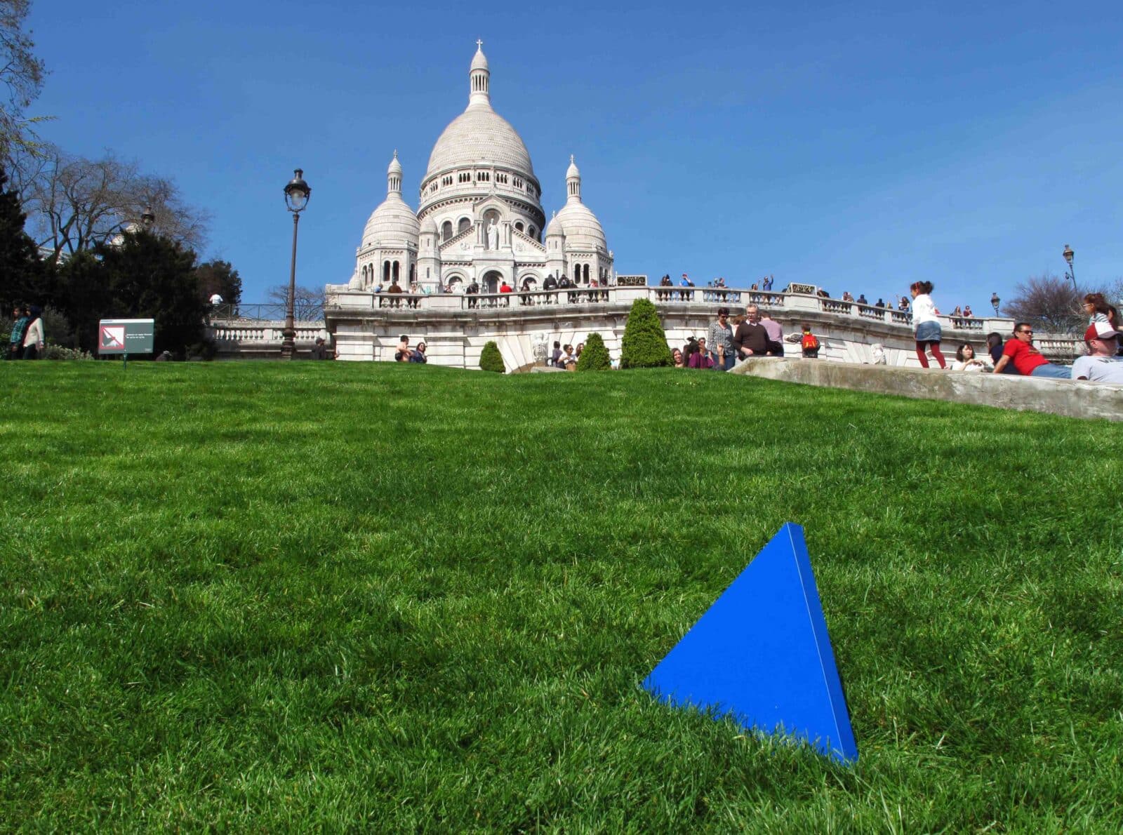 Ein grasbewachsener Hügel mit einem blauen dreieckigen Objekt im Vordergrund, Menschen, die sitzen und spazieren gehen, und die weiß gekrönte Basilika Sacré-Cœur im Hintergrund unter einem klaren blauen Himmel.