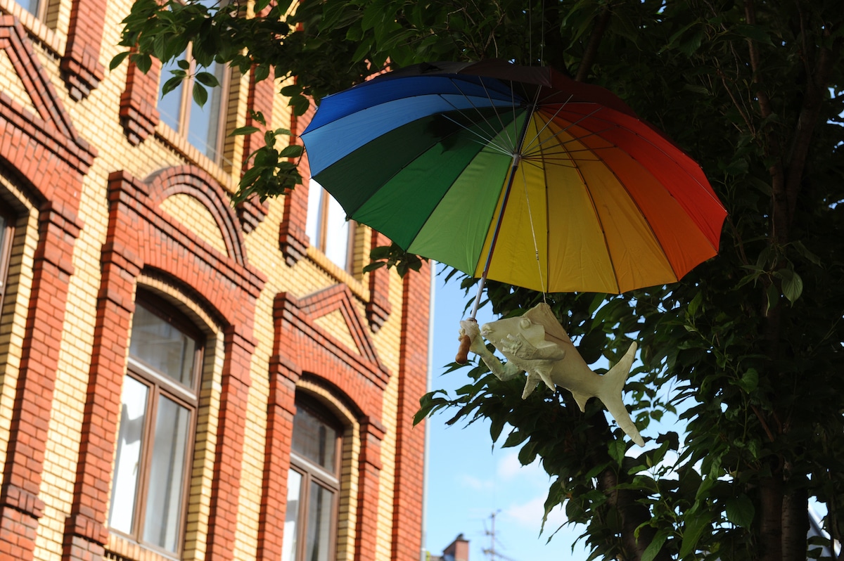 Ein bunter Regenbogenschirm hängt an einem Ast in der Nähe eines Backsteingebäudes, und unter dem Schirm hängt ein kleiner weißer Fisch.