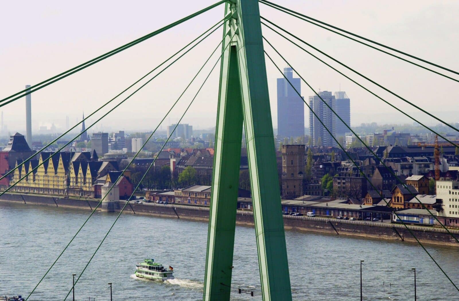 Eine große grüne Schrägseilbrücke führt über einen Fluss, unter der ein Boot hindurchfährt. Am gegenüberliegenden Ufer stehen Gebäude der Stadt, darunter moderne Hochhäuser und historische Bauten, unter einem bewölkten Himmel.
